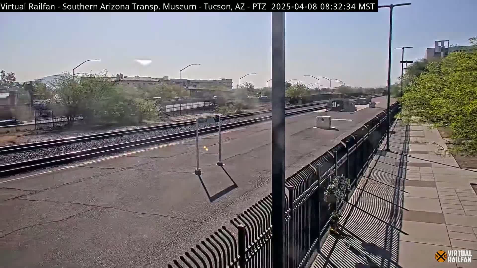 A sunny day at a train station with multiple tracks, a paved platform, and a black metal fence separating the tracks from a paved walkway.