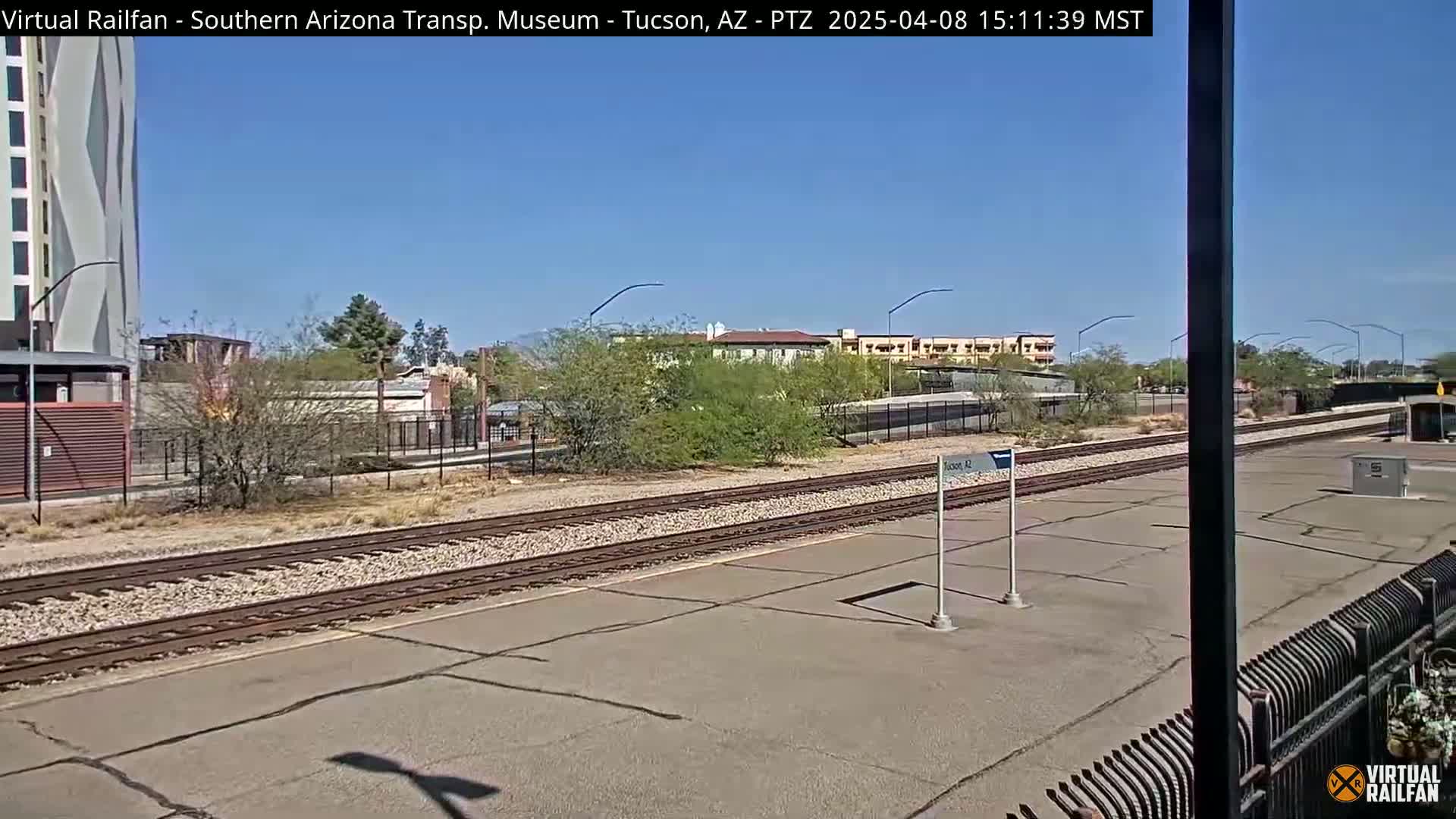 A sunny outdoor view of a train station platform with several tracks, adjacent buildings, and desert vegetation.