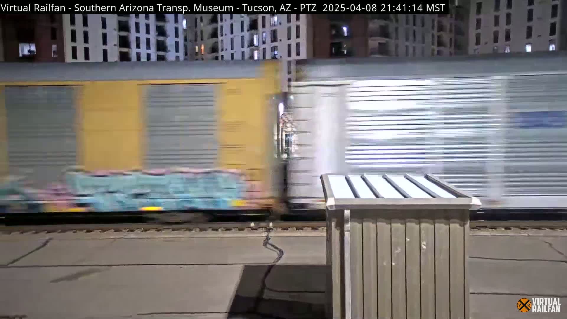A freight train with graffiti-covered cars passes by at night, near a building.