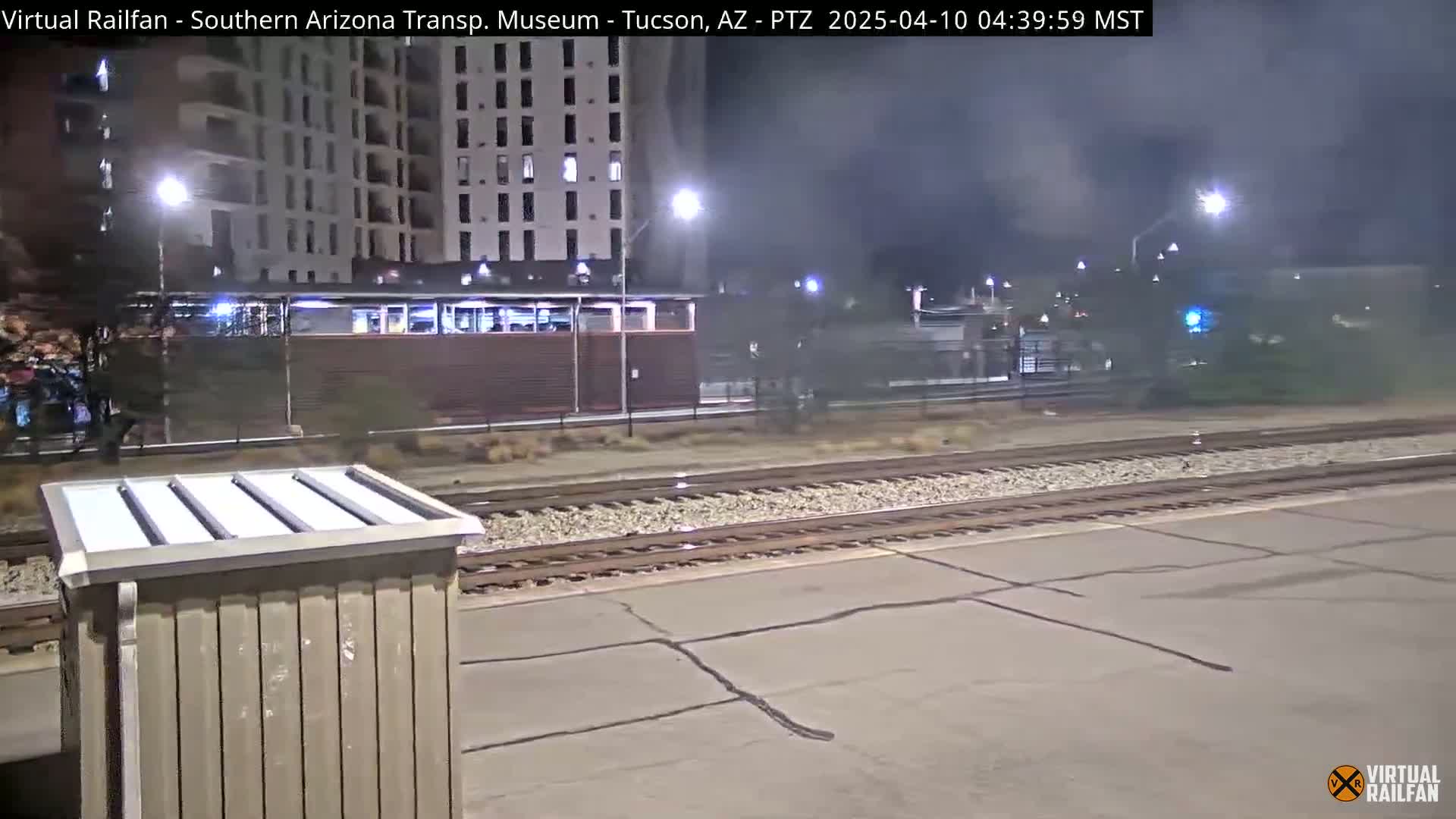 At night, a train passes by a station near a multi-story building under hazy conditions.