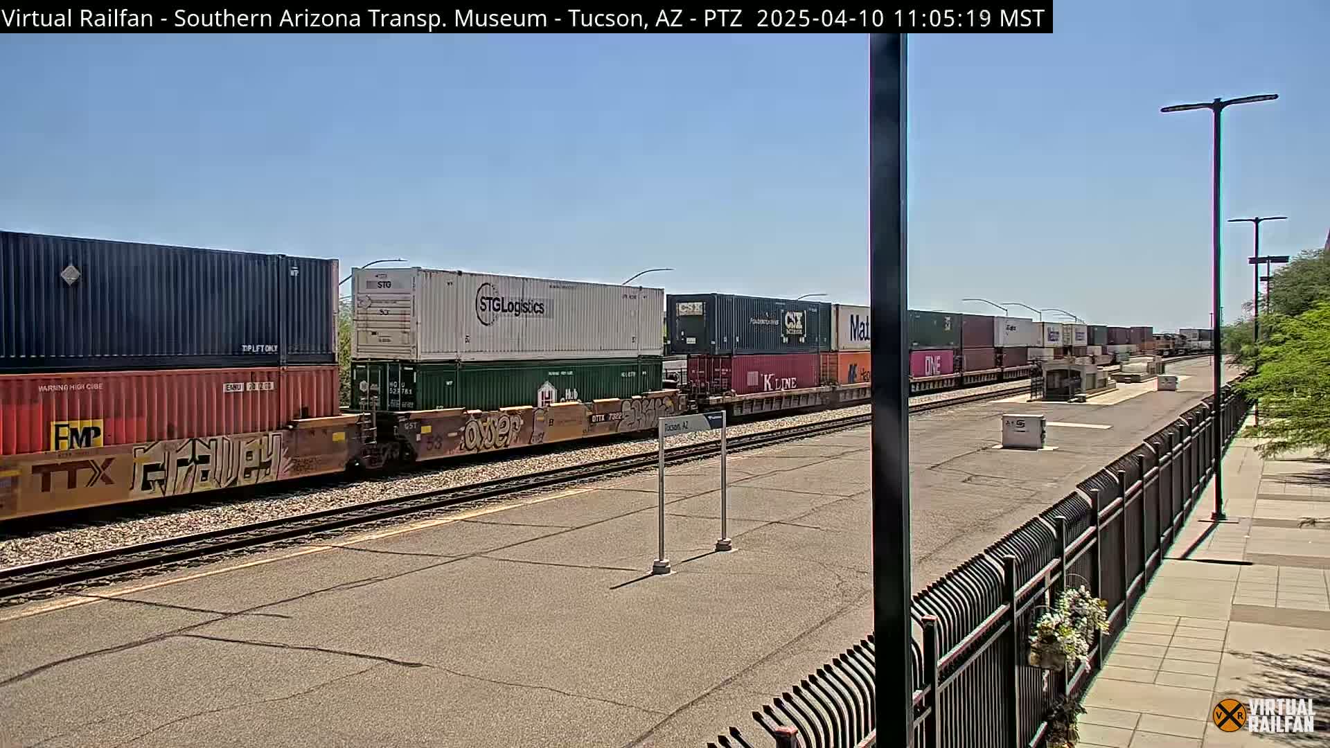 A long freight train carrying multiple shipping containers sits on a railway track under a clear, sunny sky.