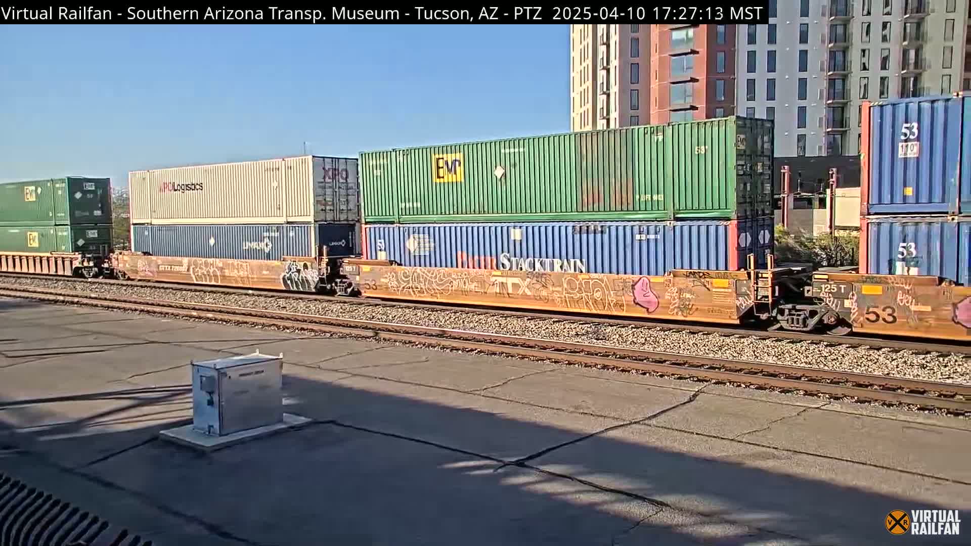 A freight train carrying multiple shipping containers moves along railroad tracks on a sunny day, passing near modern buildings.