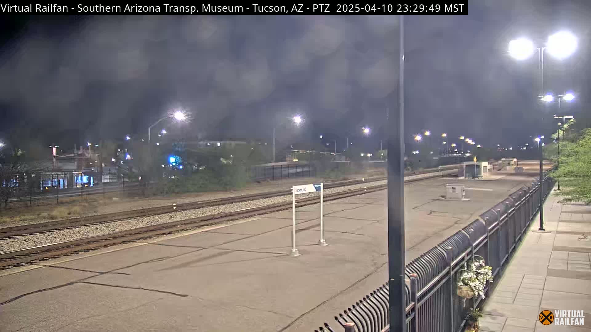 A nighttime view of a mostly empty train station platform, under a hazy, dark sky, with train tracks extending into the distance.