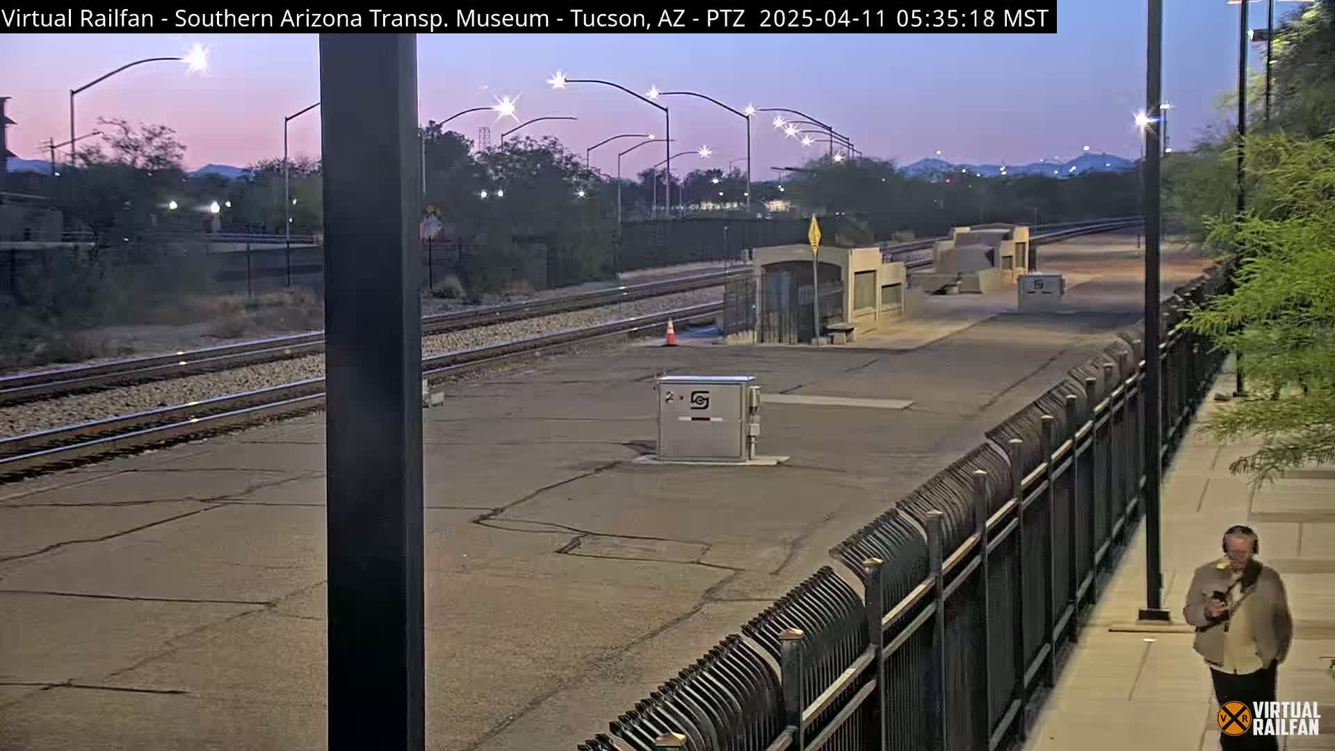 A deserted train station platform at dawn, under a twilight sky, with a single person walking along a sidewalk beside a fence.