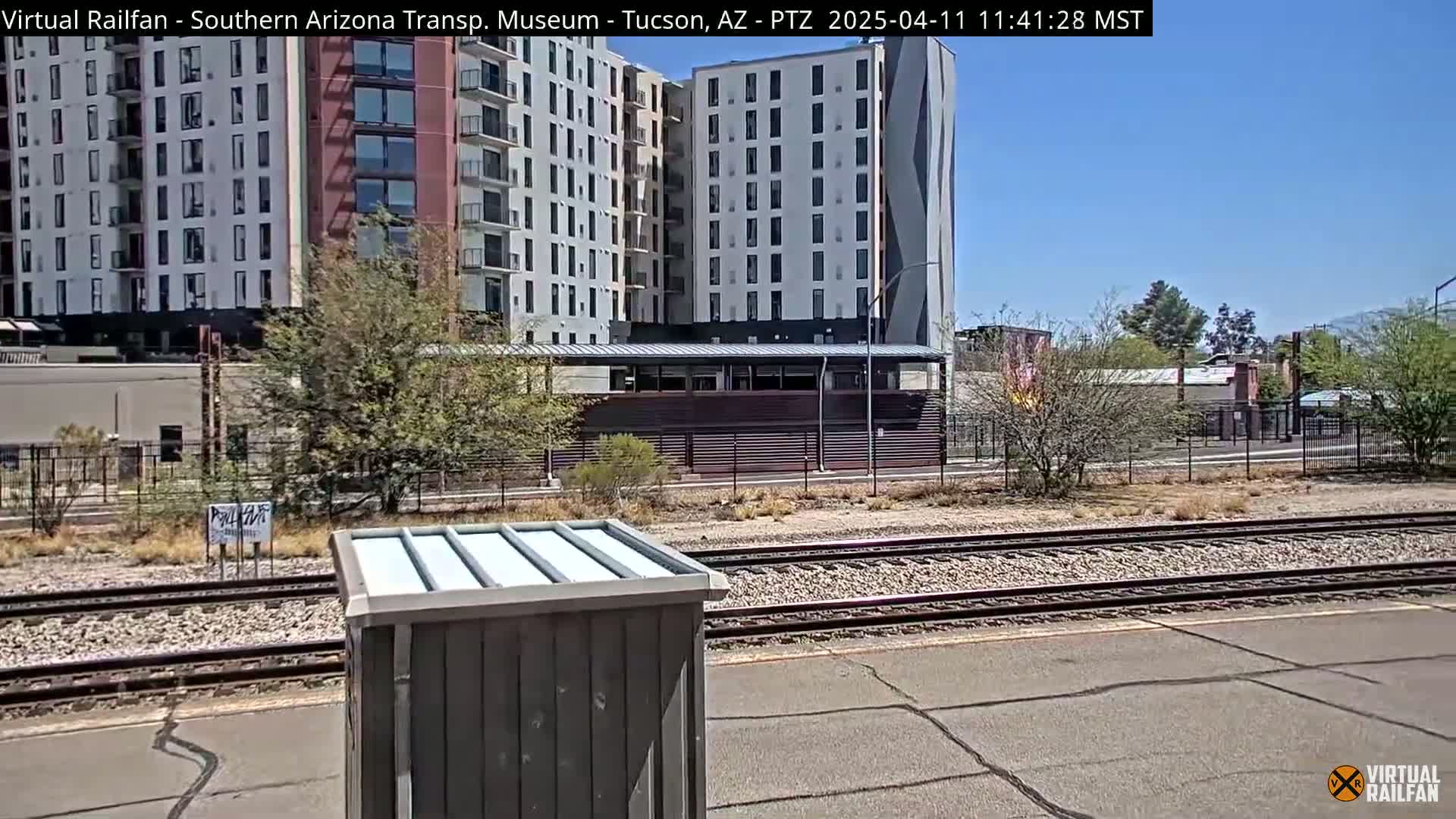 A view of railroad tracks on a sunny day, with a tall apartment building and some scrubby trees in the background.