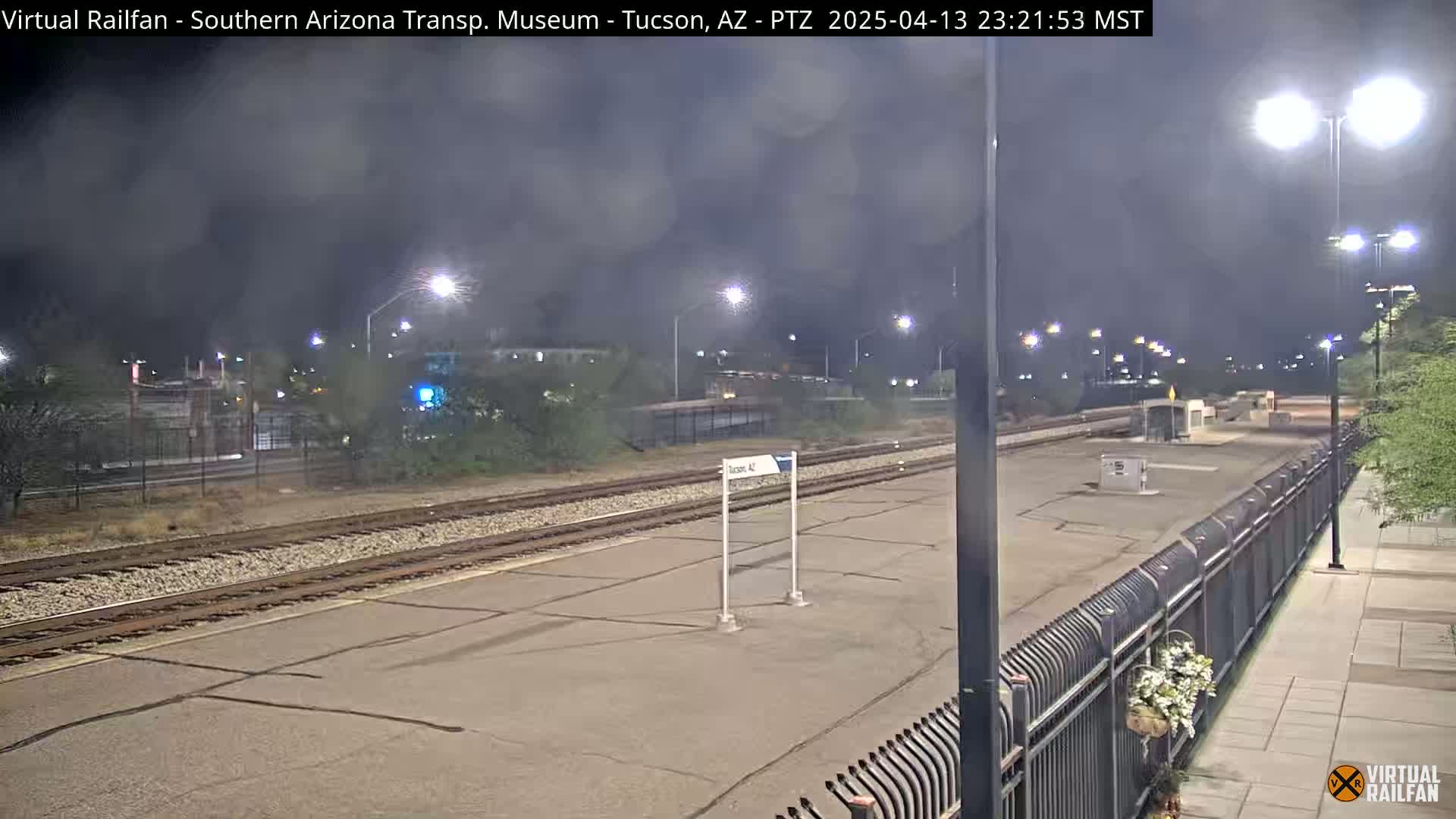 A nighttime view of a deserted train station platform with railroad tracks, illuminated by streetlights under a cloudy sky.