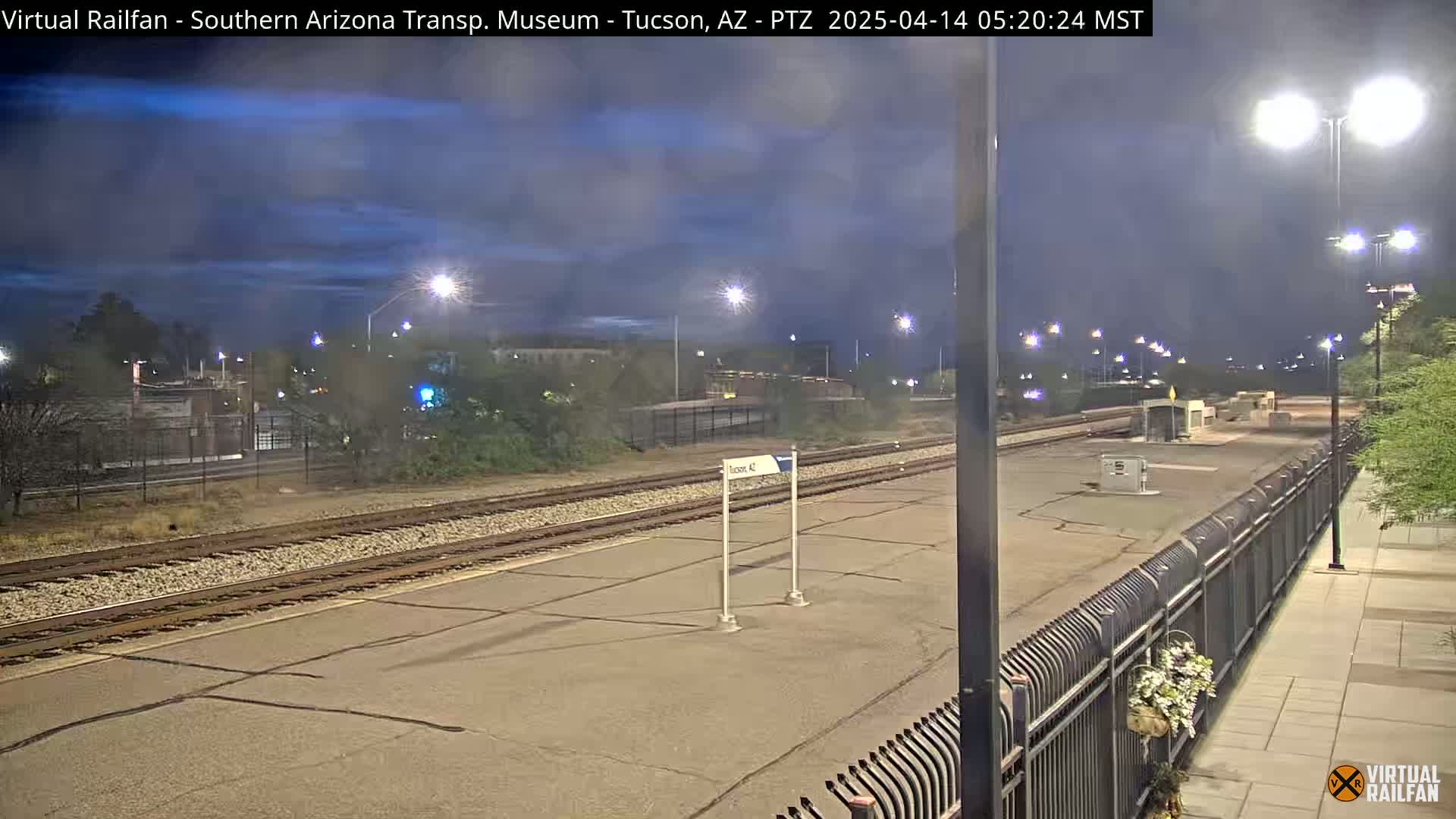 A nighttime view of a train station platform with train tracks, streetlights, and some vegetation under a twilight sky.