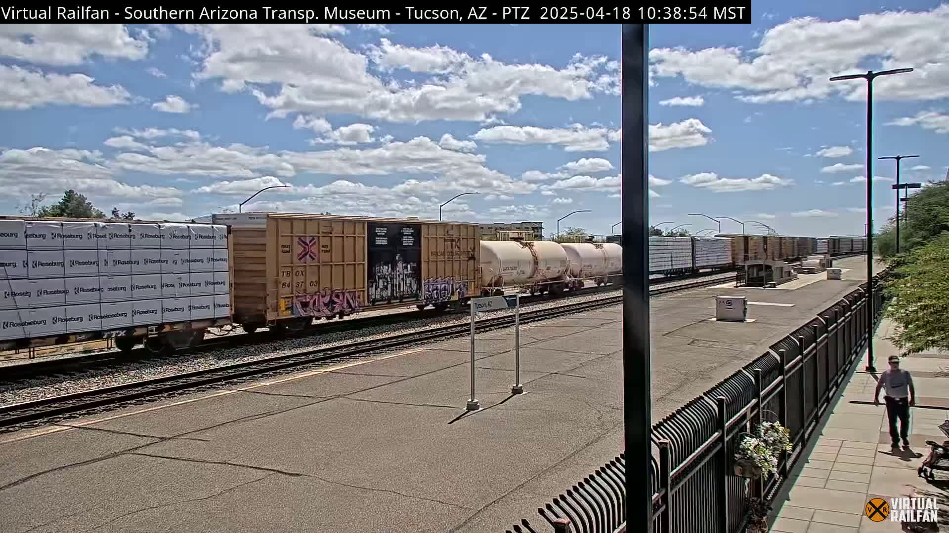 A partly cloudy day shows a freight train carrying lumber and tanker cars sitting on a track next to a paved area with a person walking by a fence.