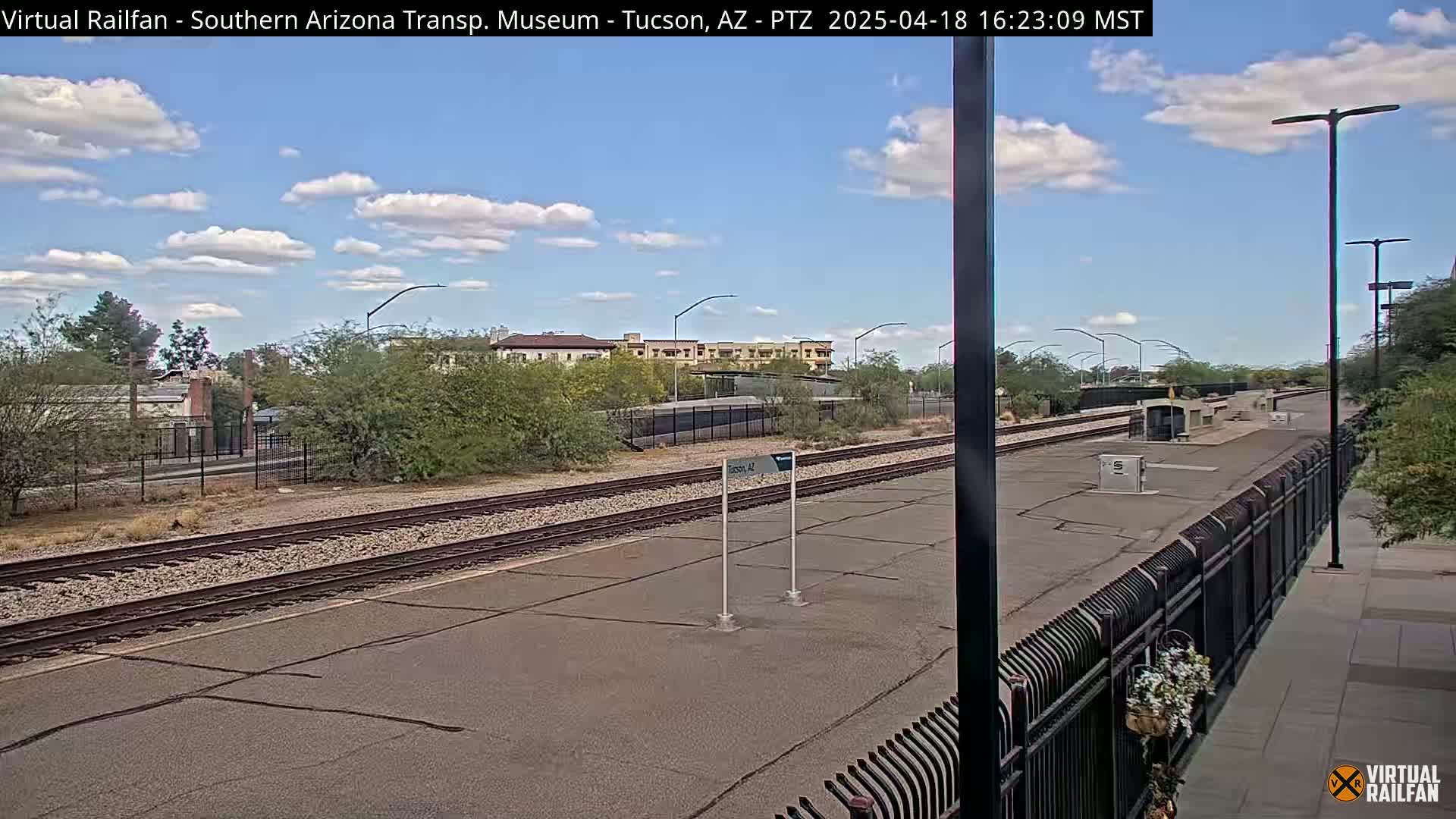 A mostly sunny day at a train station with a train visible in the distance, featuring a paved platform, railroad tracks, and desert landscaping.