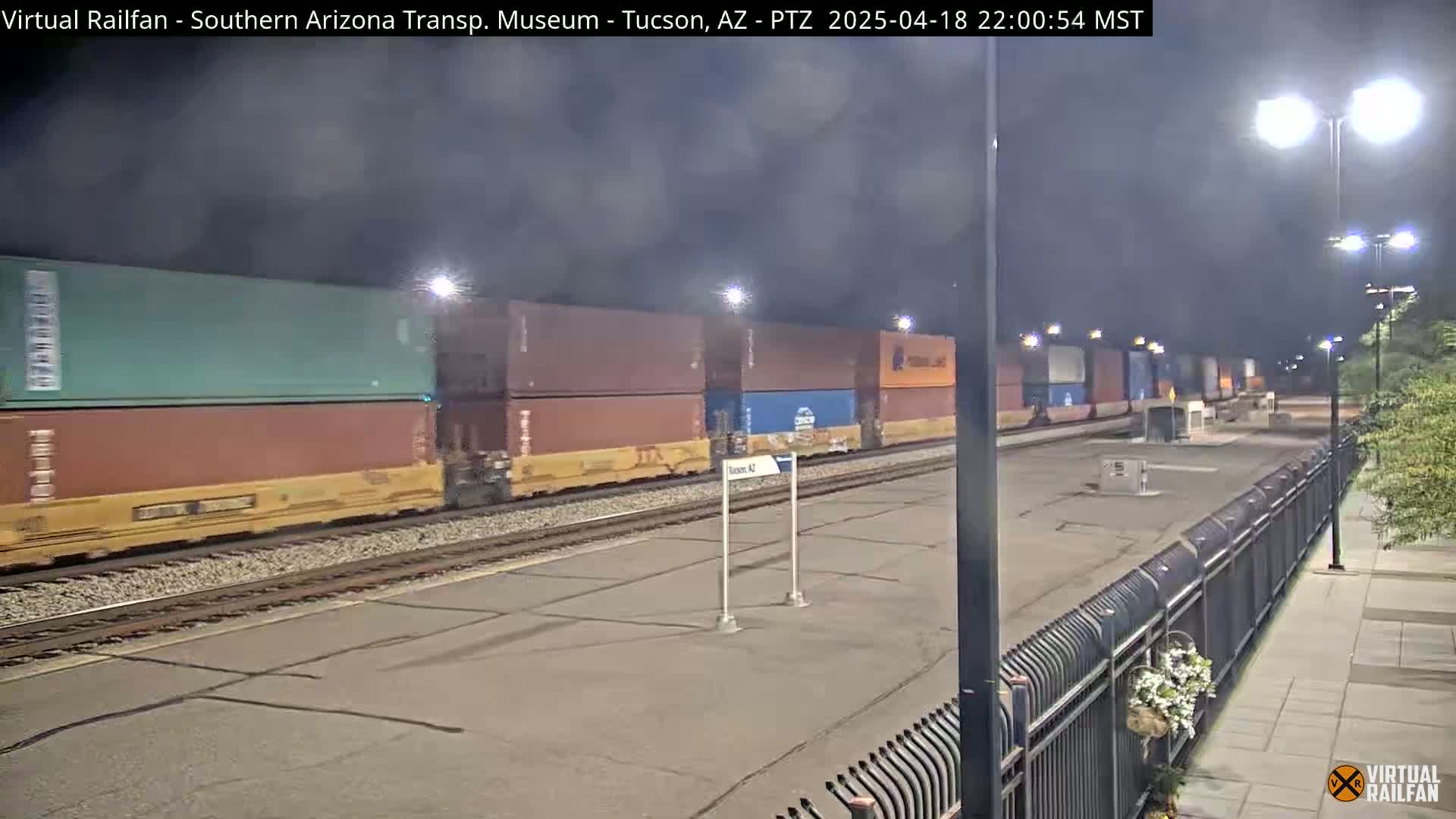 A long freight train carrying stacked shipping containers moves slowly along railway tracks at night under hazy, dark skies.
