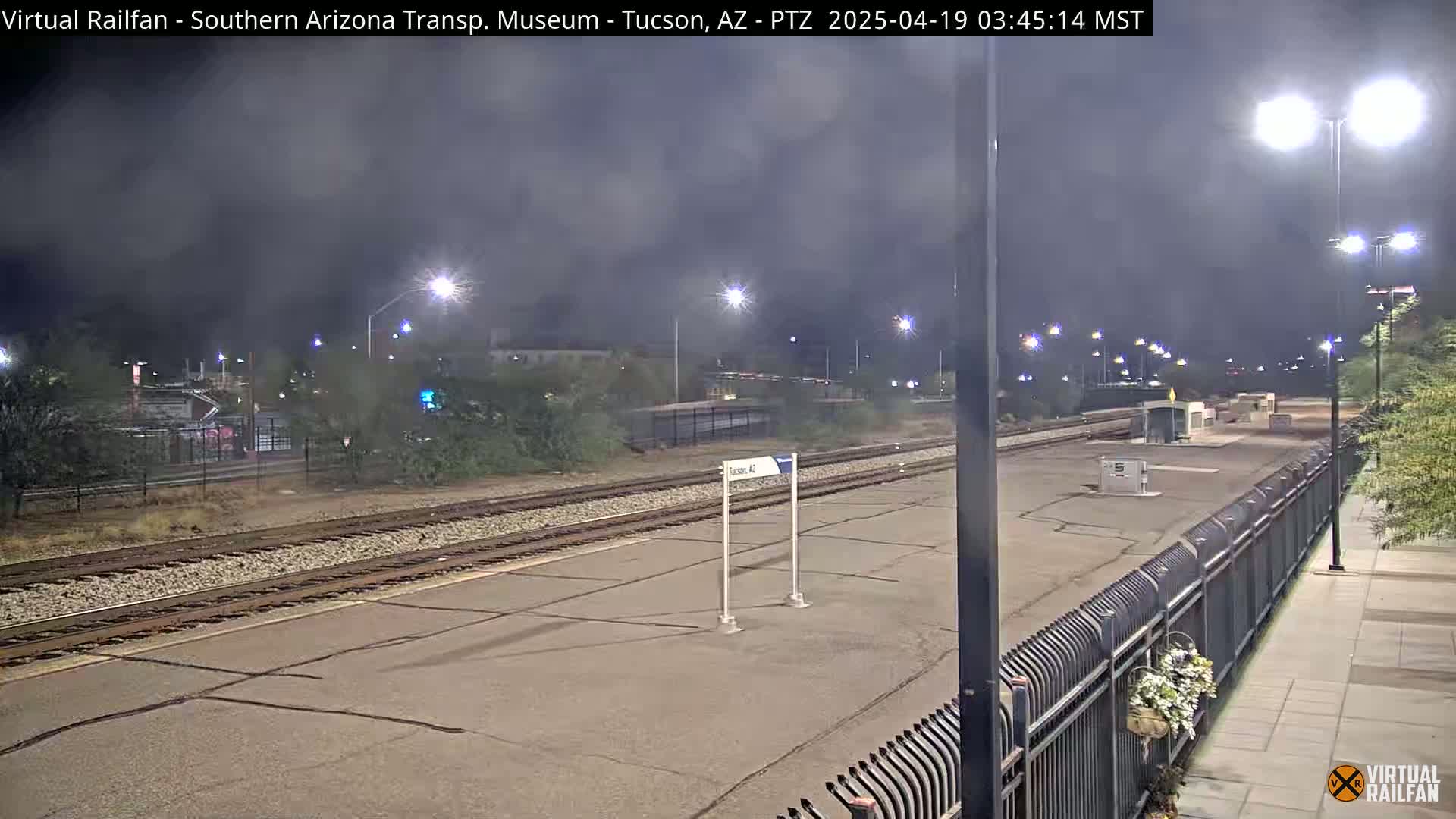 A nighttime view of a train platform with a hazy sky, illuminated by streetlights, and a metal fence separating the platform from a paved walkway.
