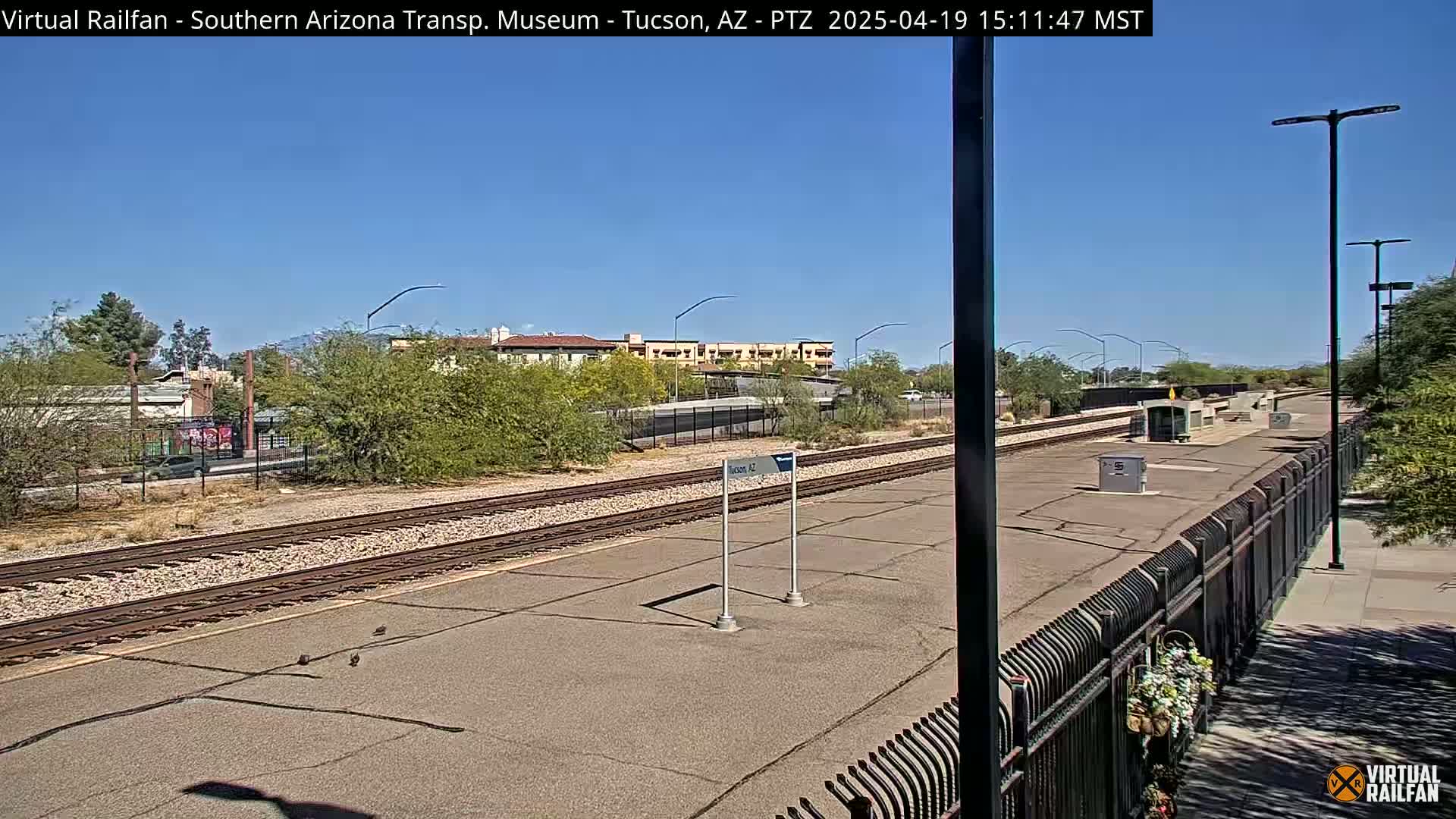 A train station platform with a clear blue sky and several tracks is shown, surrounded by desert landscaping and buildings.
