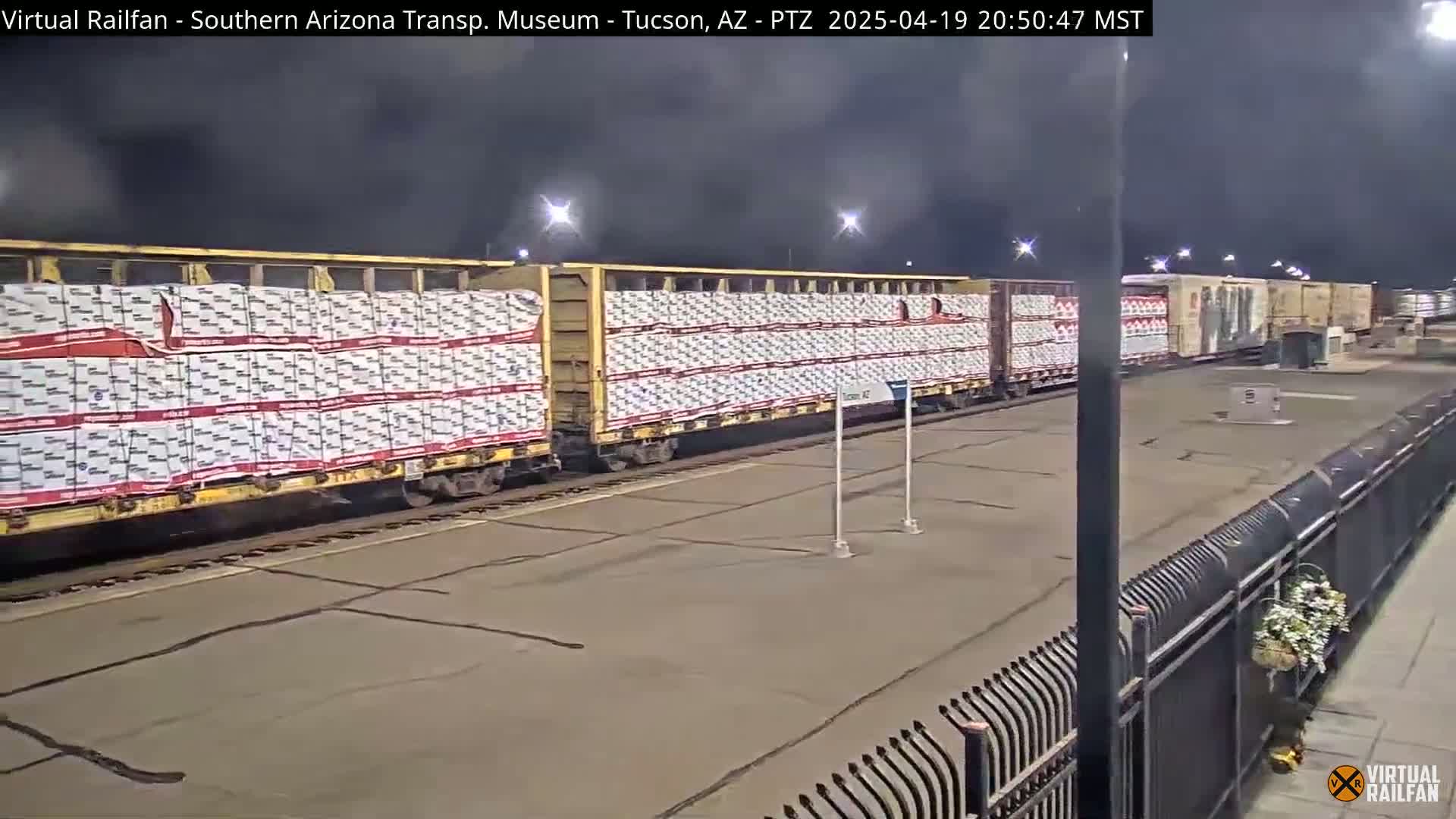 A long freight train, loaded with uniformly packaged cargo, sits stationary under a dark night sky illuminated by artificial lights.