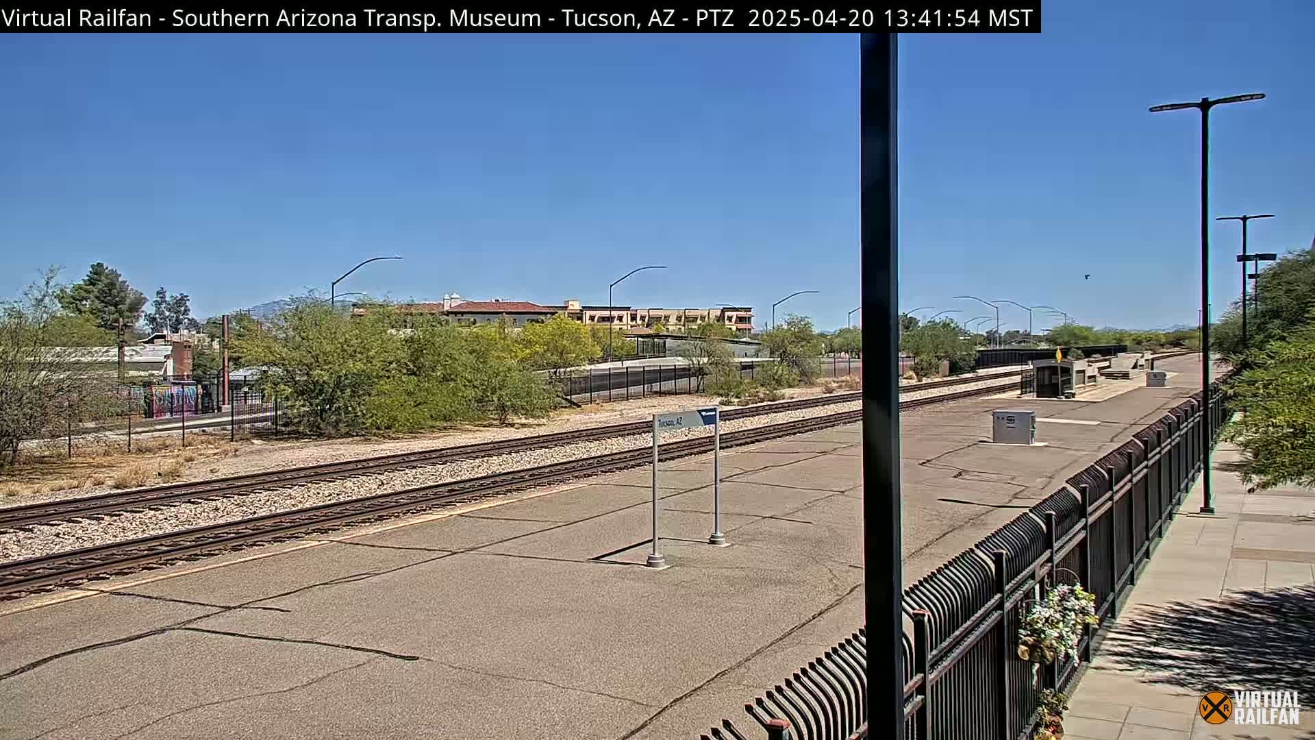 A sunny day reveals a train platform with railroad tracks extending into the distance, bordered by a black metal fence, and surrounded by desert landscaping and buildings.