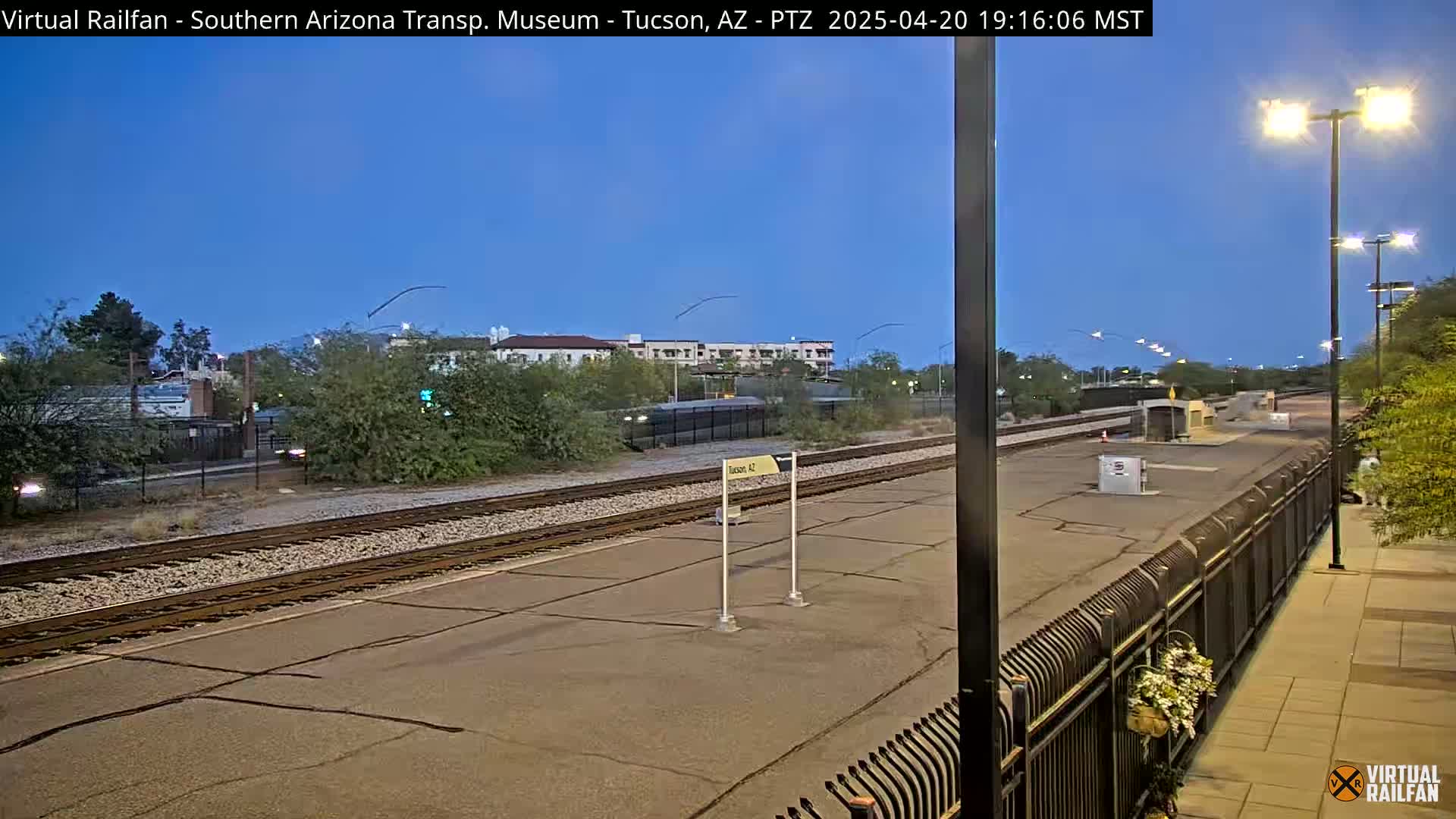 A train platform at dusk, under a clear twilight sky, features train tracks extending into the distance, with a building visible in the background.