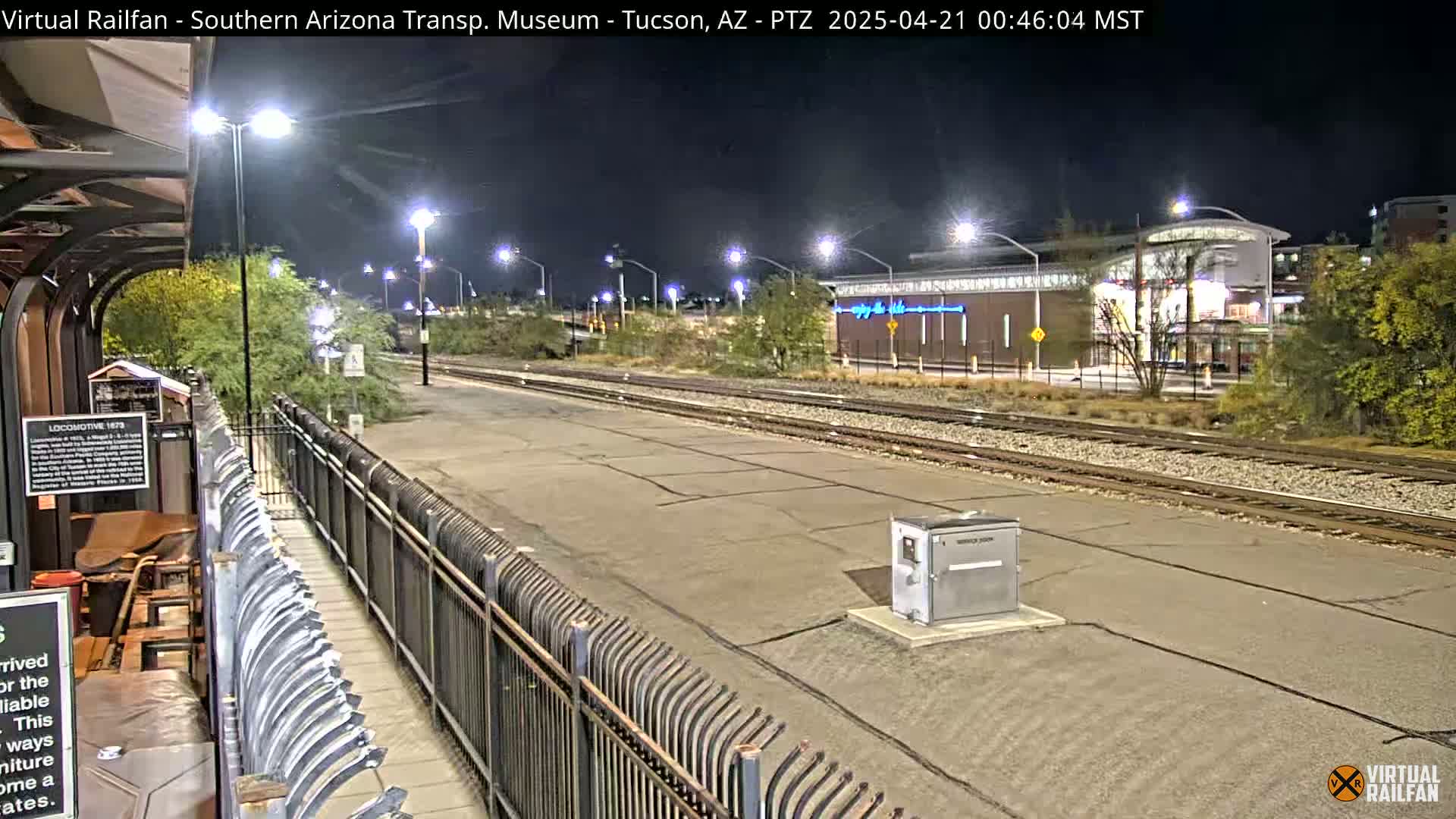 A nighttime view of a train station platform and tracks, illuminated by lights, with a building in the background.