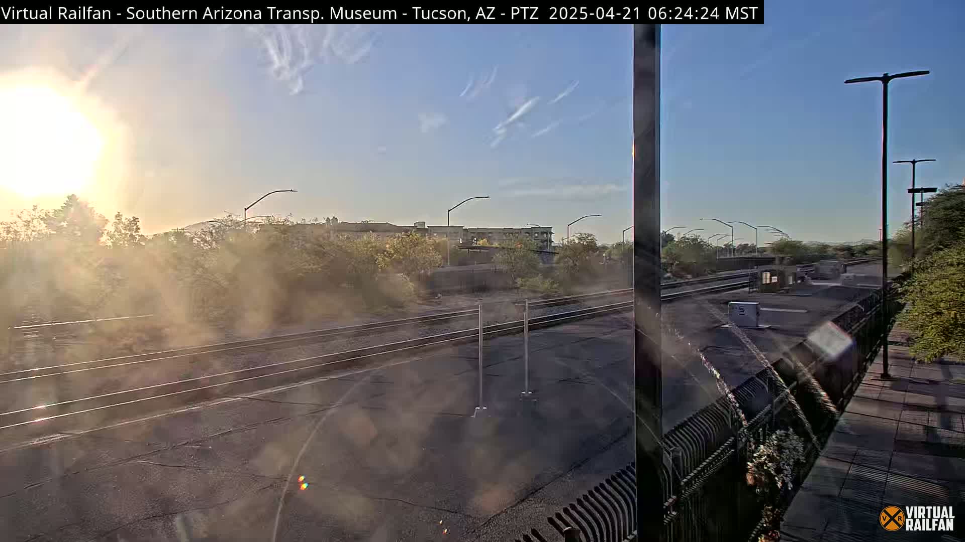 A sunny morning view of train tracks and a station platform, with low vegetation and buildings in the background.