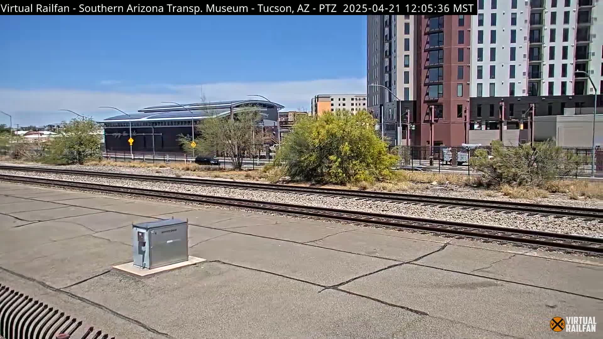 A sunny day shows railroad tracks beside a paved area, with a building and a tall apartment building in the background, and desert vegetation in between.