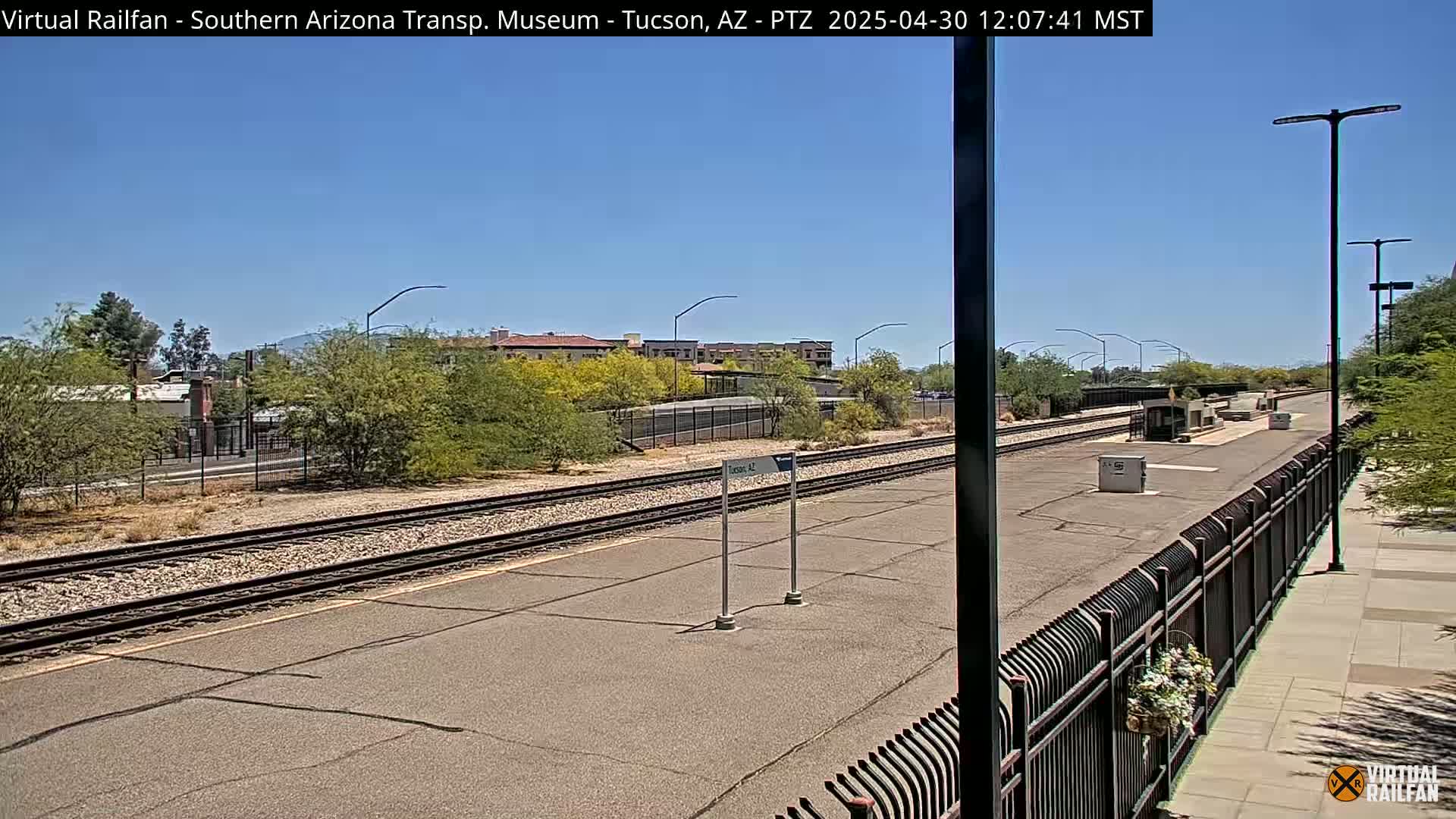 A sunny day at a train station with railroad tracks stretching into the distance, bordered by desert landscaping and buildings.