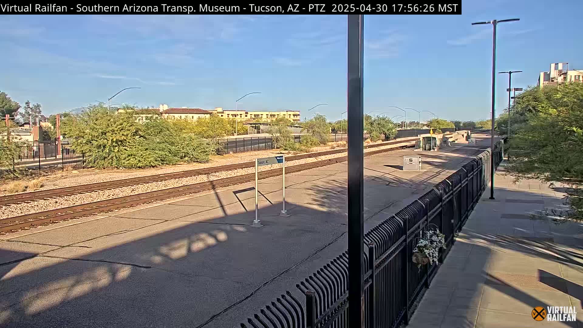 A sunny day shows a train station platform with train tracks, a fence, and desert vegetation, with buildings visible in the background.