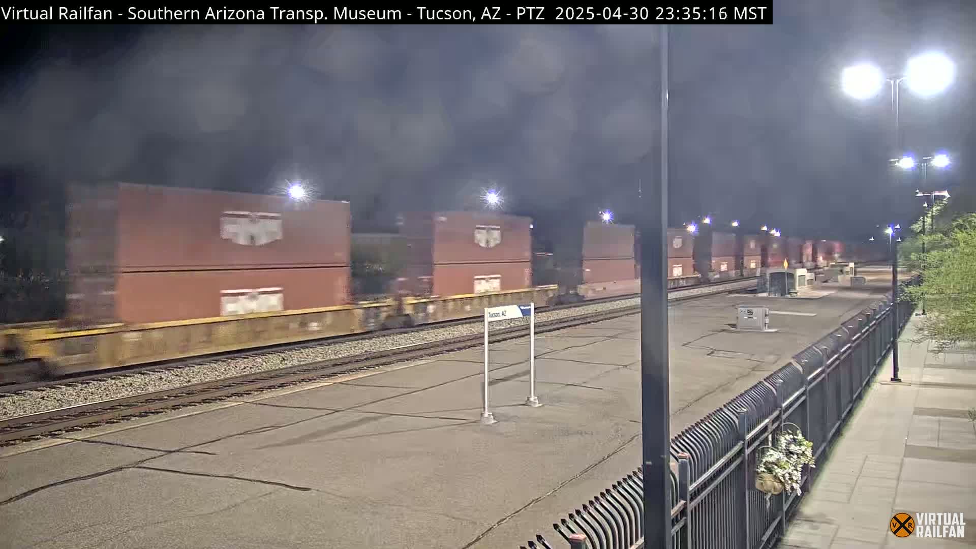 A long freight train carrying numerous shipping containers moves past a station at night under dark, cloudy skies.