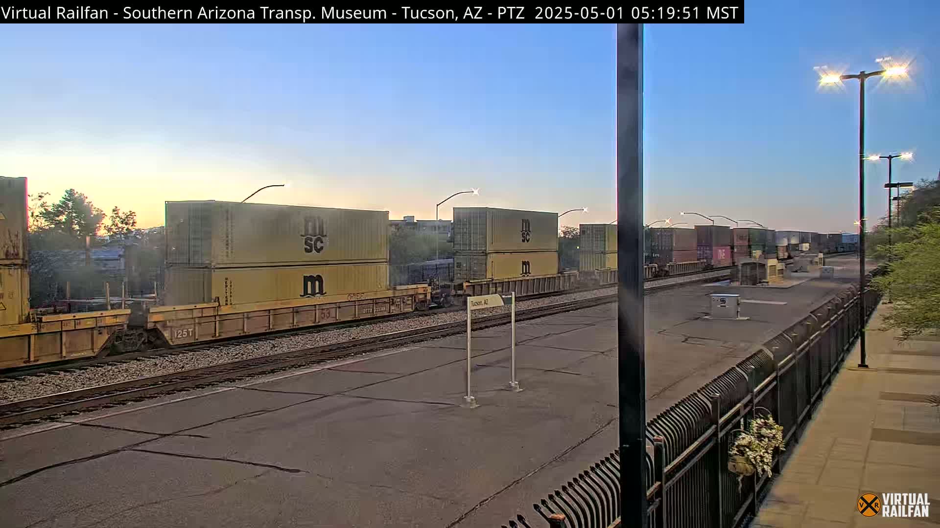 A long freight train carrying stacked shipping containers sits on a track at sunrise under a clear sky.