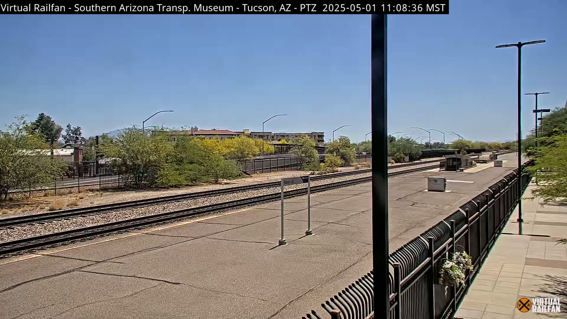 A sunny day at a train station with a platform, tracks, and shrubbery,  under a clear blue sky.