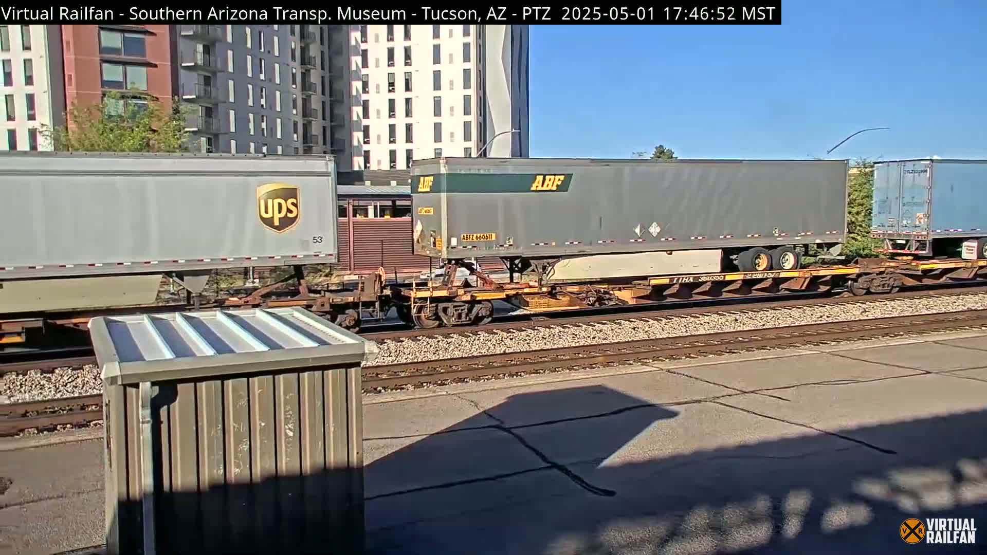 A freight train carrying several shipping containers, including one marked UPS and another marked ABF, sits on a rail line under a clear, sunny sky near an apartment building.