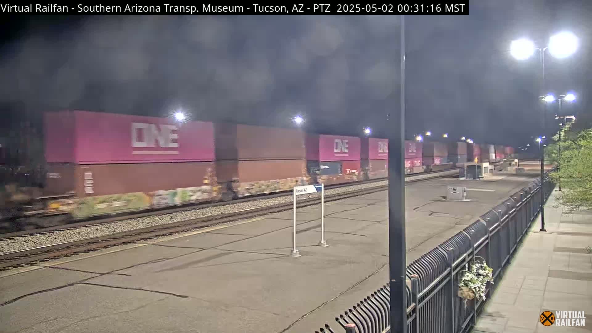 A long freight train with pink containers passes by a brightly lit train station at night.