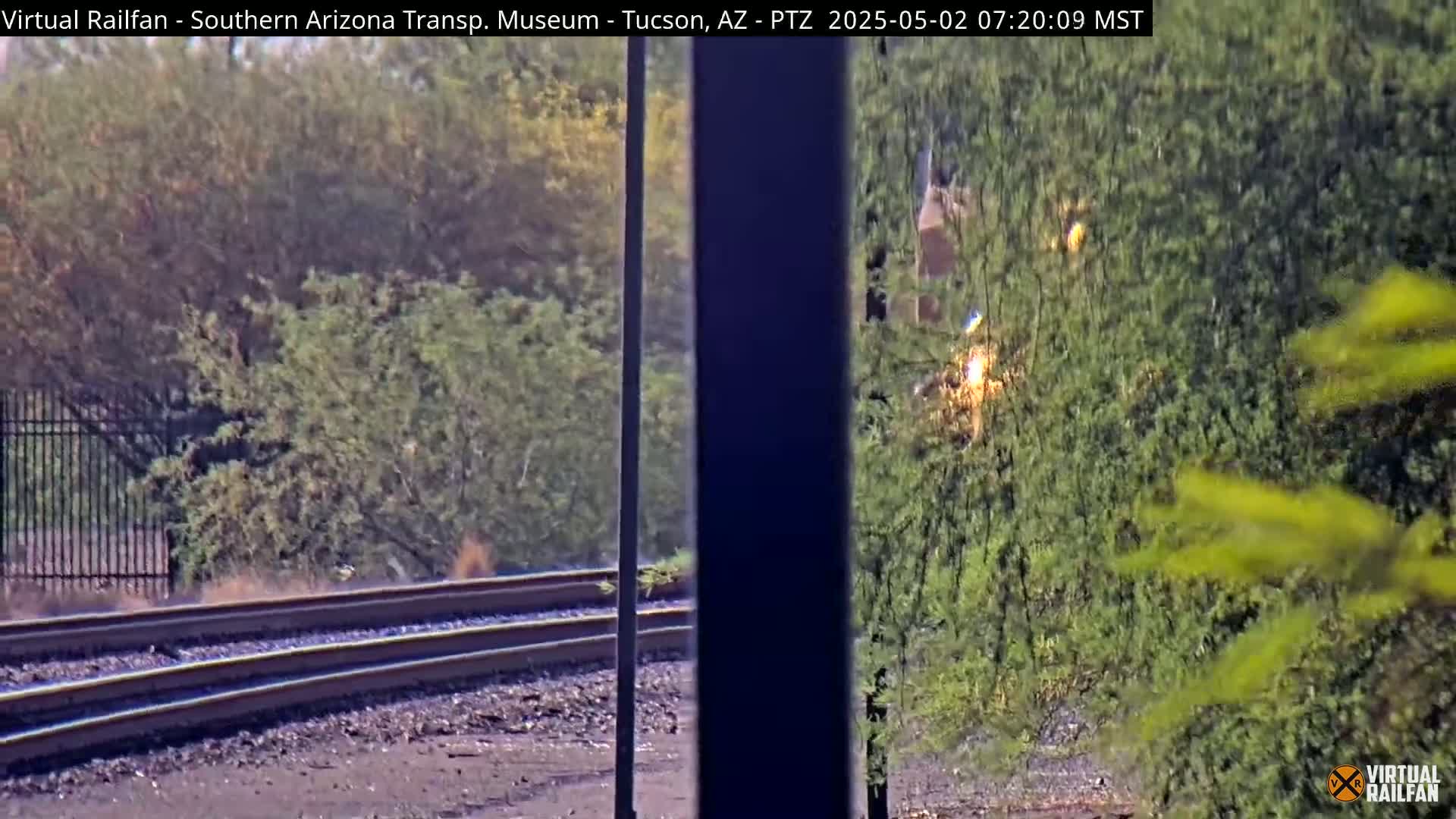 A train passes behind a partially obscured view of railroad tracks and lush green vegetation on a sunny day.