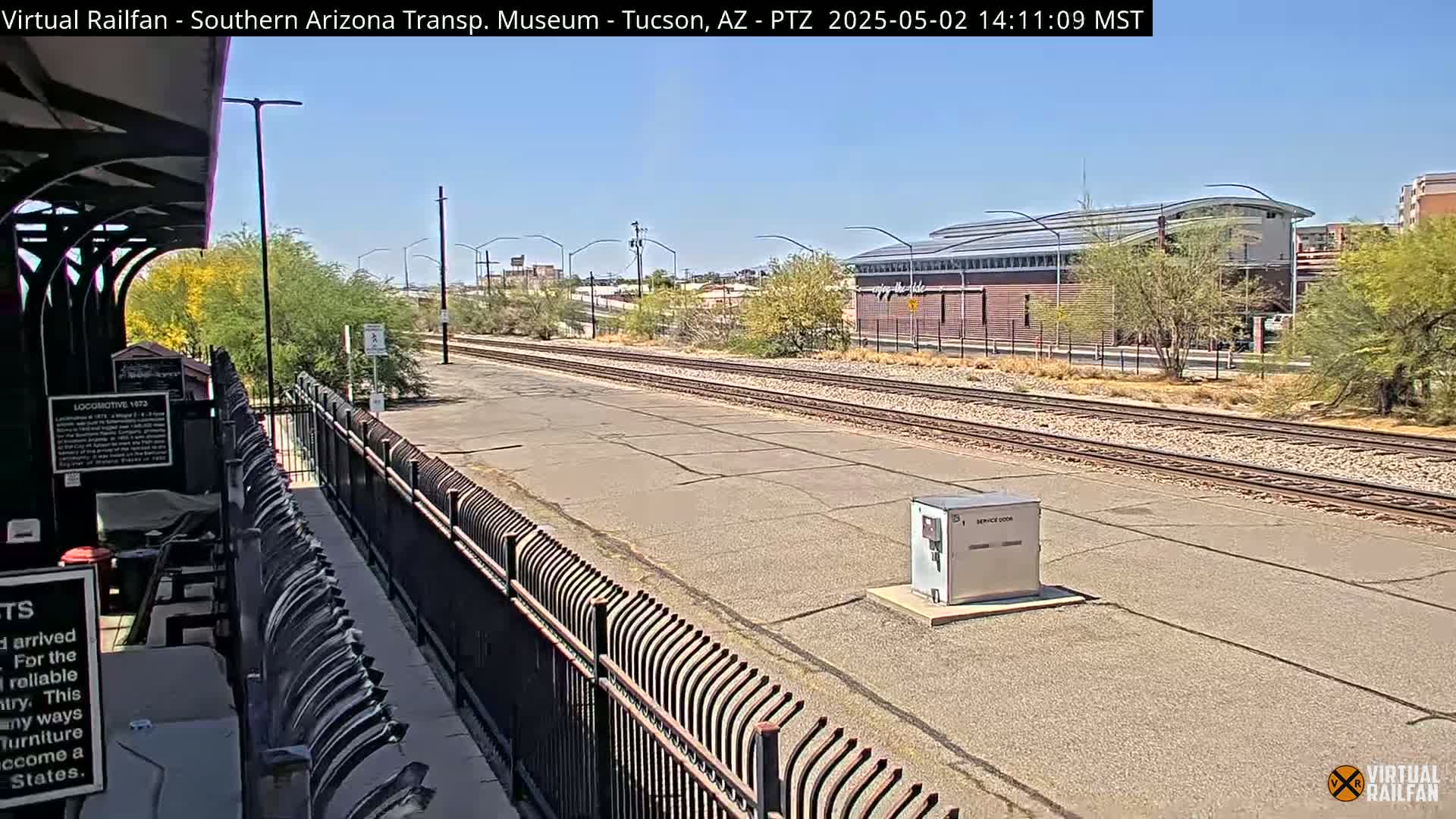 A sunny day at a train station shows train tracks extending into the distance past a modern building and a black metal fence.