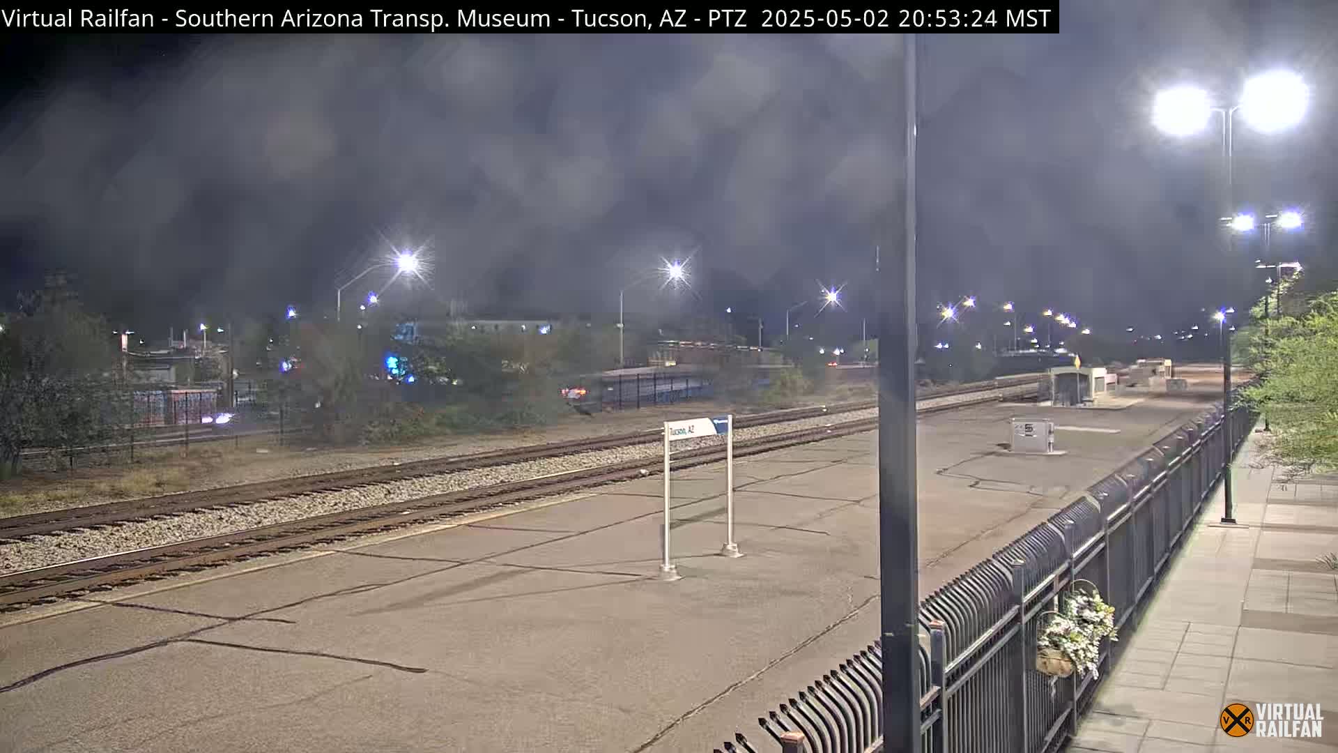 A nighttime view of a train station platform with train tracks and a hazy, dark sky.