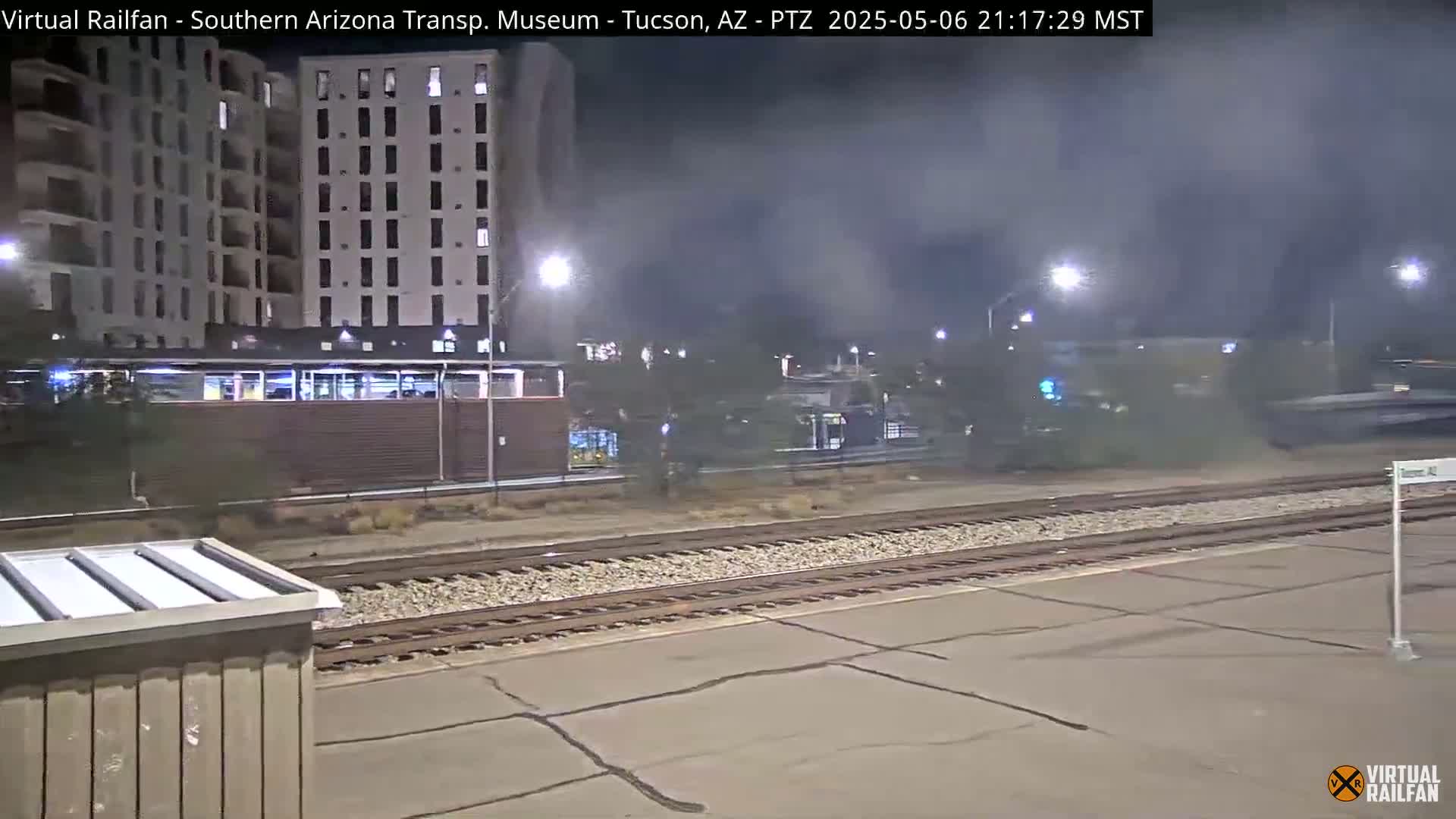 A nighttime view of a train passing by a station, near a tall building, with hazy atmospheric conditions.