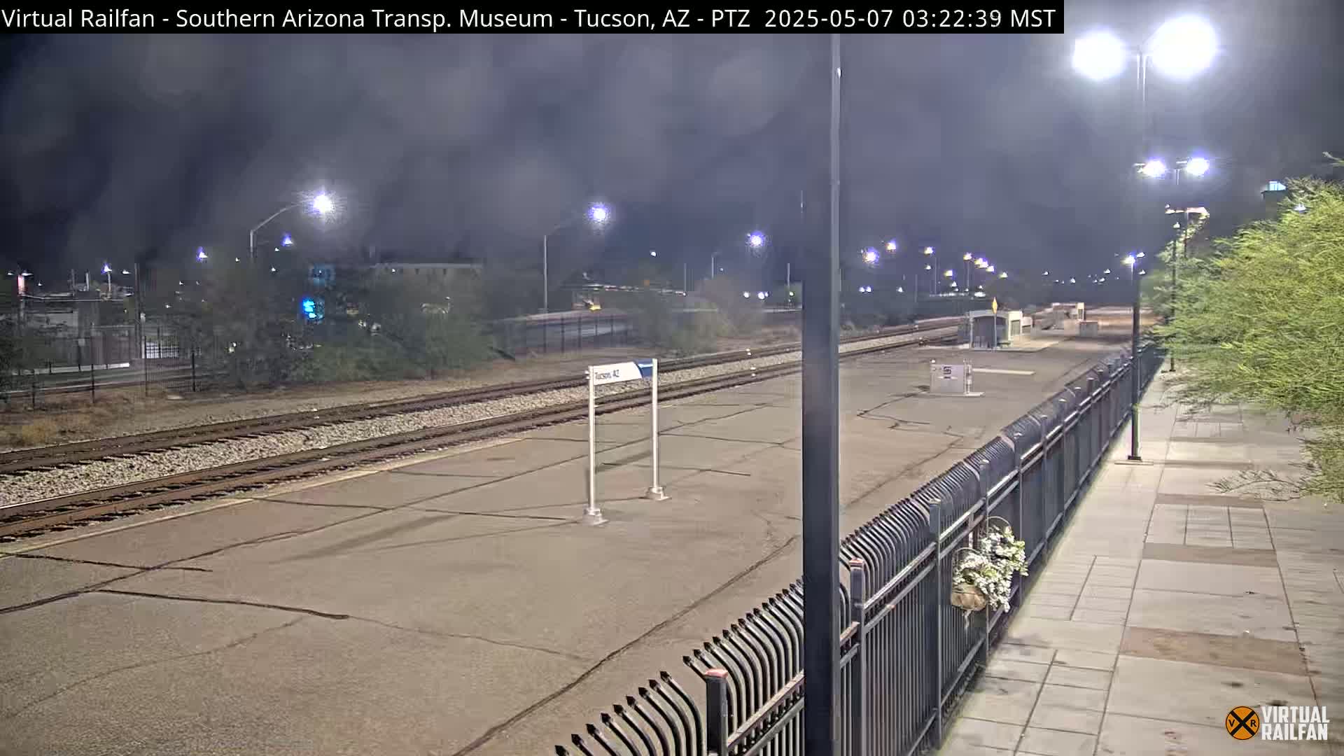 A nighttime view of a deserted train platform with multiple sets of train tracks, illuminated by streetlights, under a dark, cloudy sky.