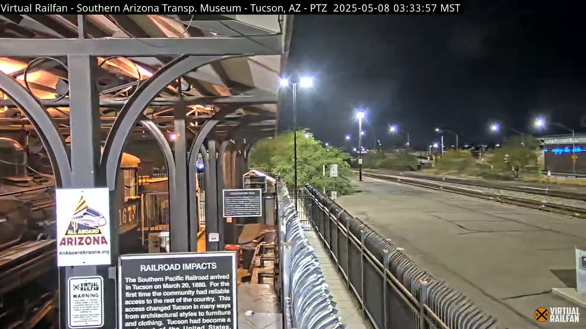 A nighttime view of a train station platform and tracks, with a metal fence and sparse desert vegetation visible.