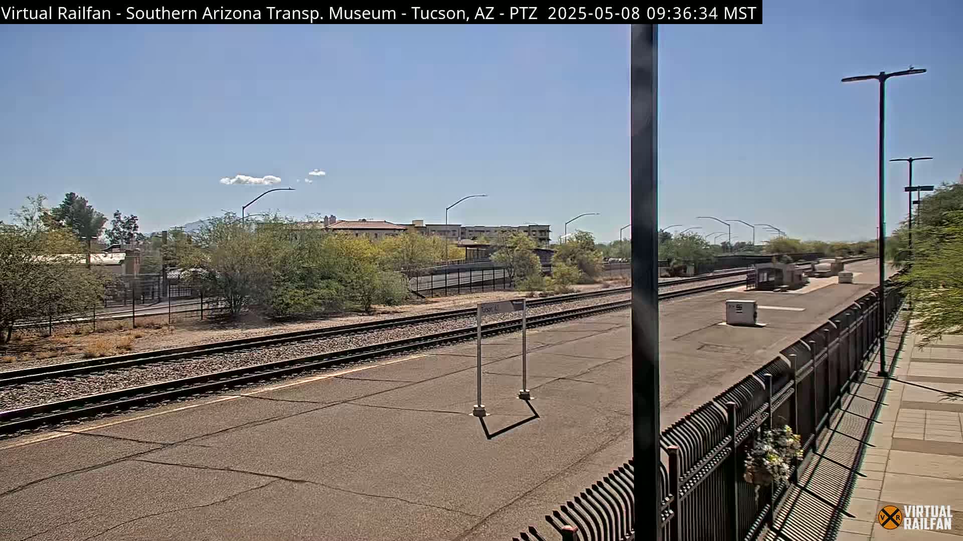 A sunny day at a train station with two sets of train tracks, a platform, and buildings in the background.