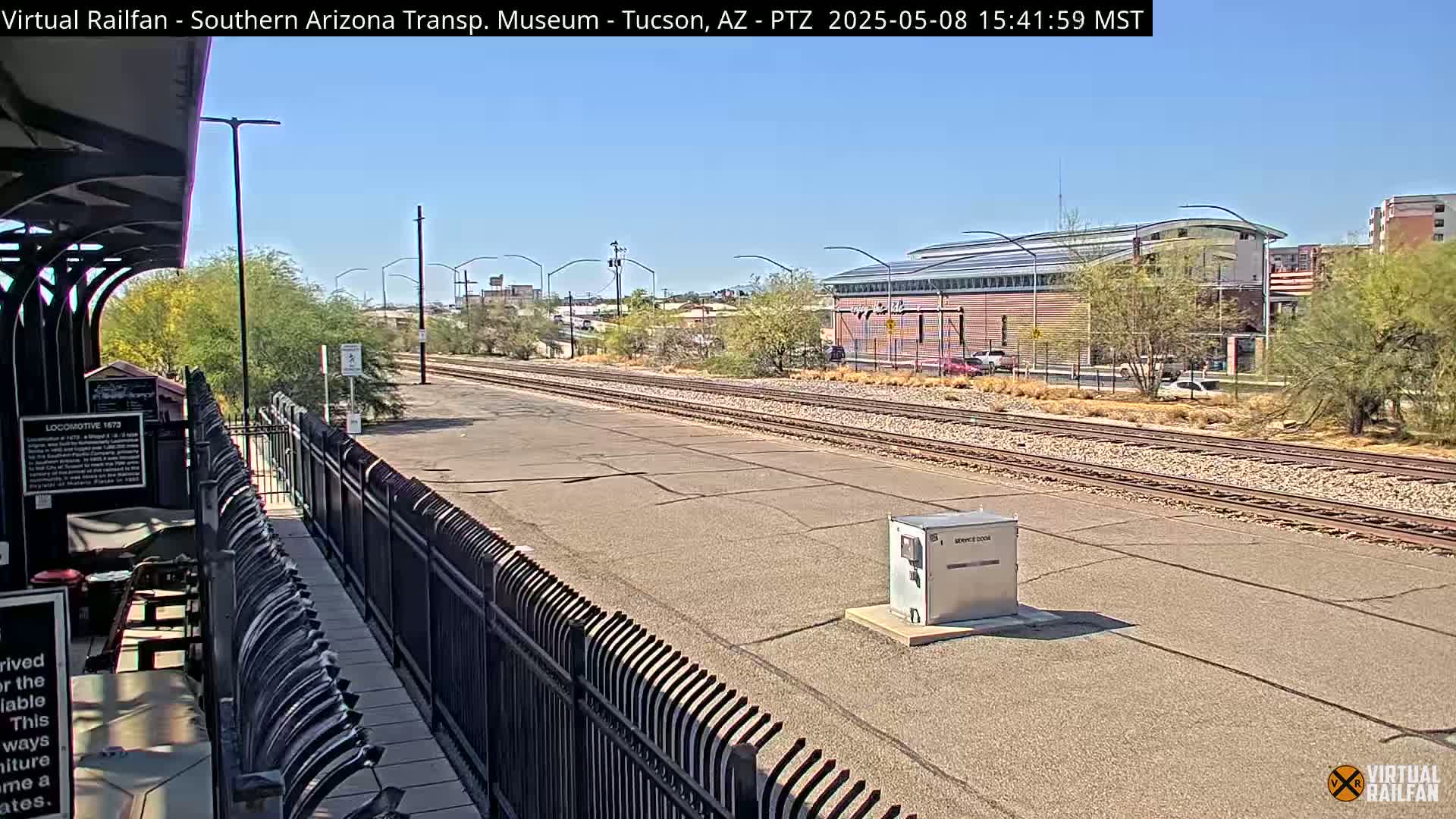 A sunny day at a train station shows railway tracks extending into the distance, passing in front of a light-brown building and several desert plants.