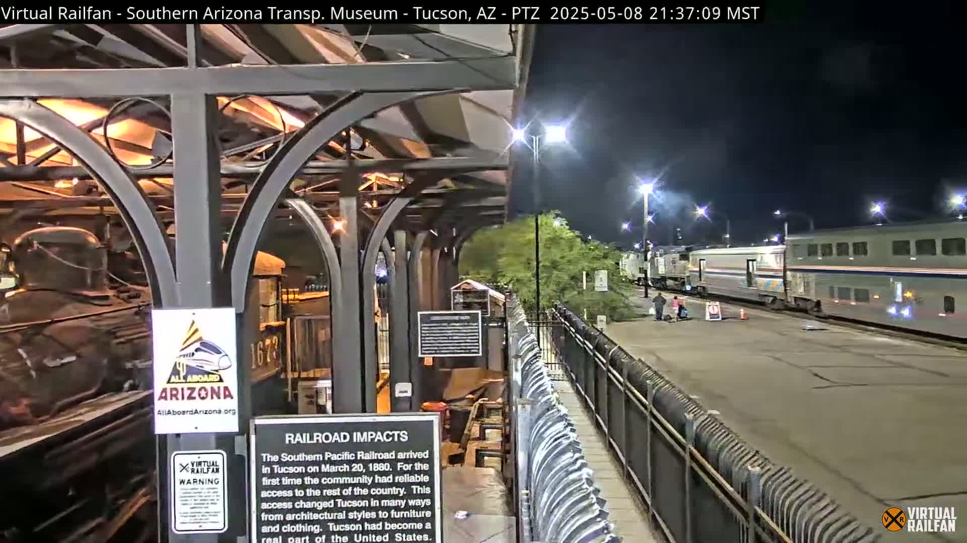 A nighttime view of a train station platform with a portion of a passenger train and a historic locomotive visible, under bright artificial lighting.