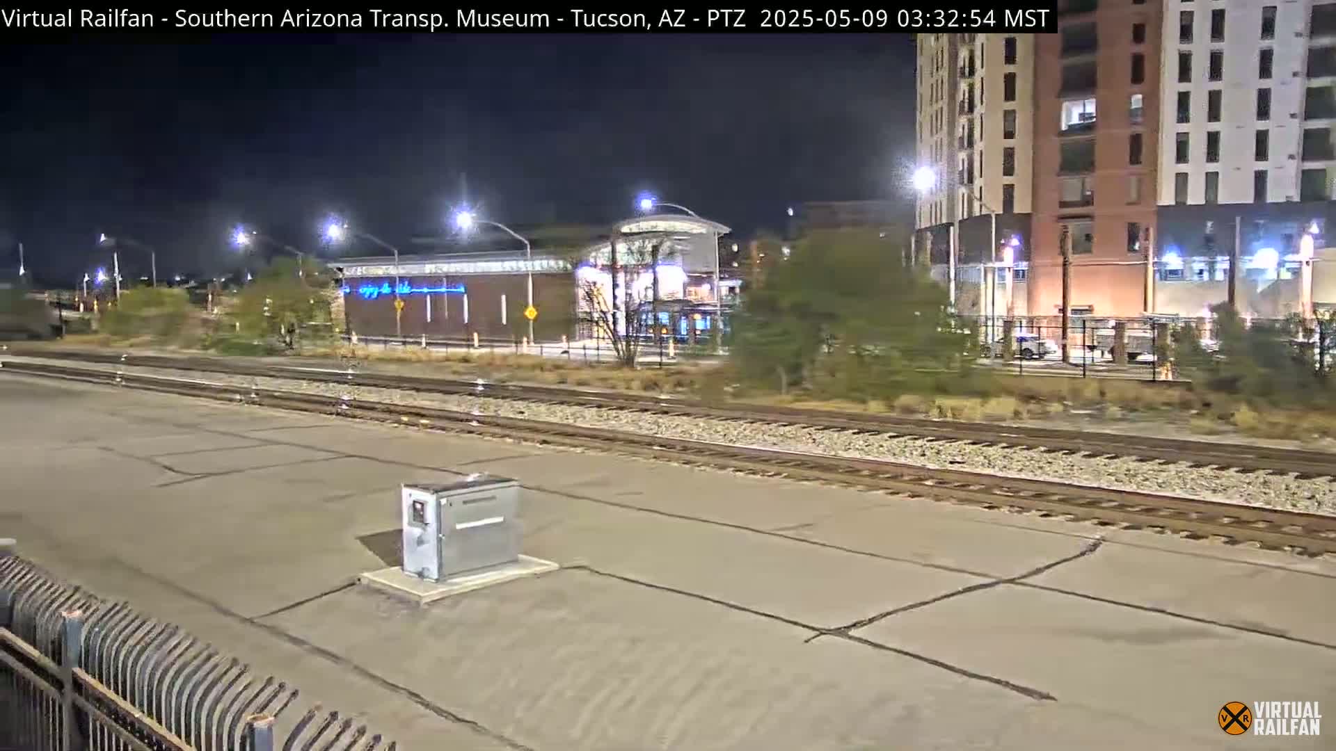 A nighttime view of train tracks at a station, with a building and streetlights visible in the background under a clear, dark sky.