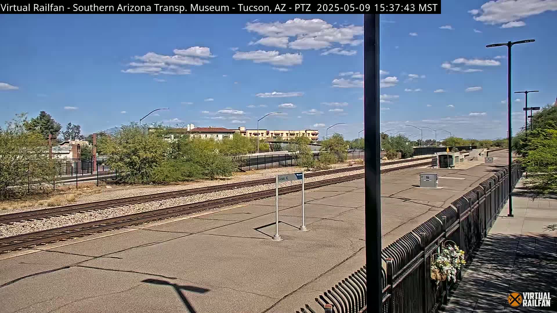 A mostly sunny day at a train station with a paved platform, train tracks, and desert landscaping.