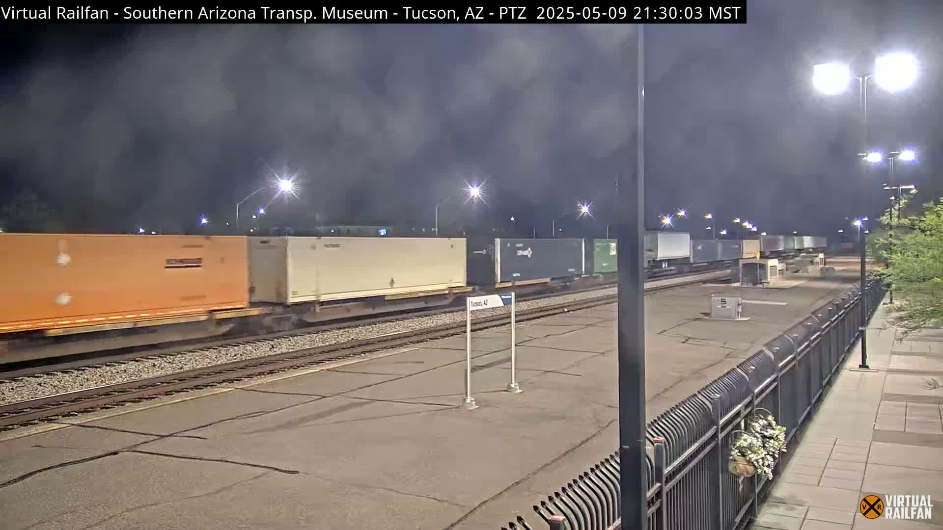 A freight train with multiple multicolored shipping containers passes by a nighttime railway station under a cloudy sky.