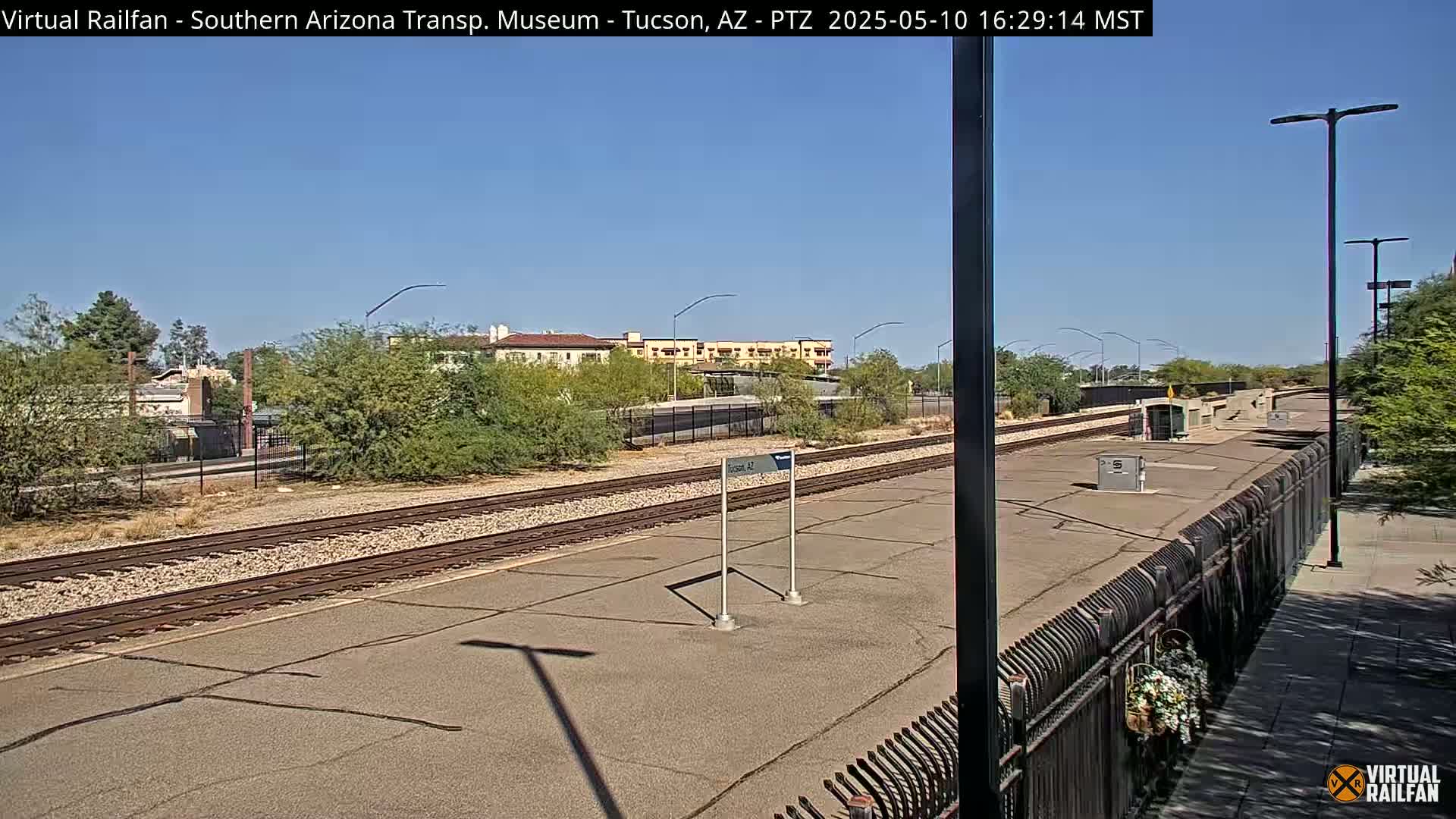 A sunny day at a train station platform with a view of train tracks, desert landscaping, and a building in the distance.