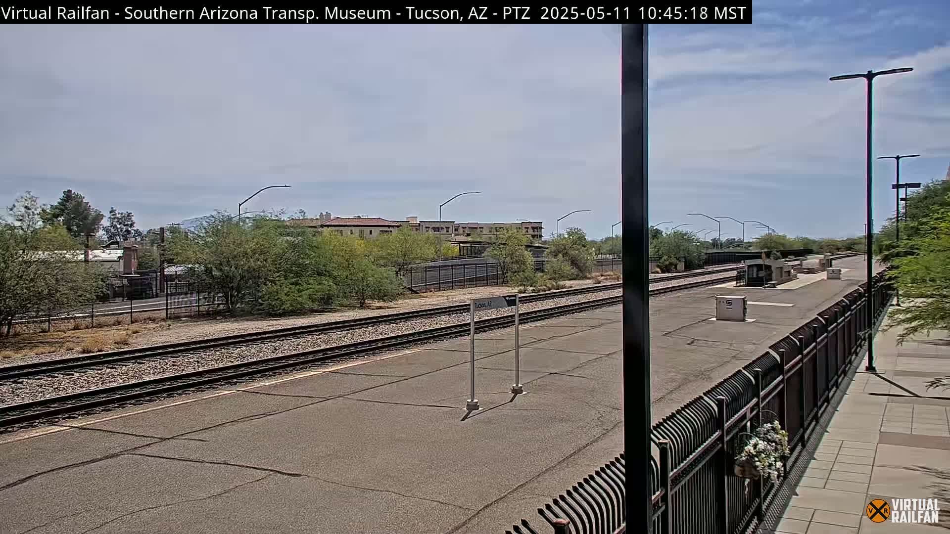 A mostly sunny day at a train station platform with multiple train tracks, a low fence, and buildings in the background.