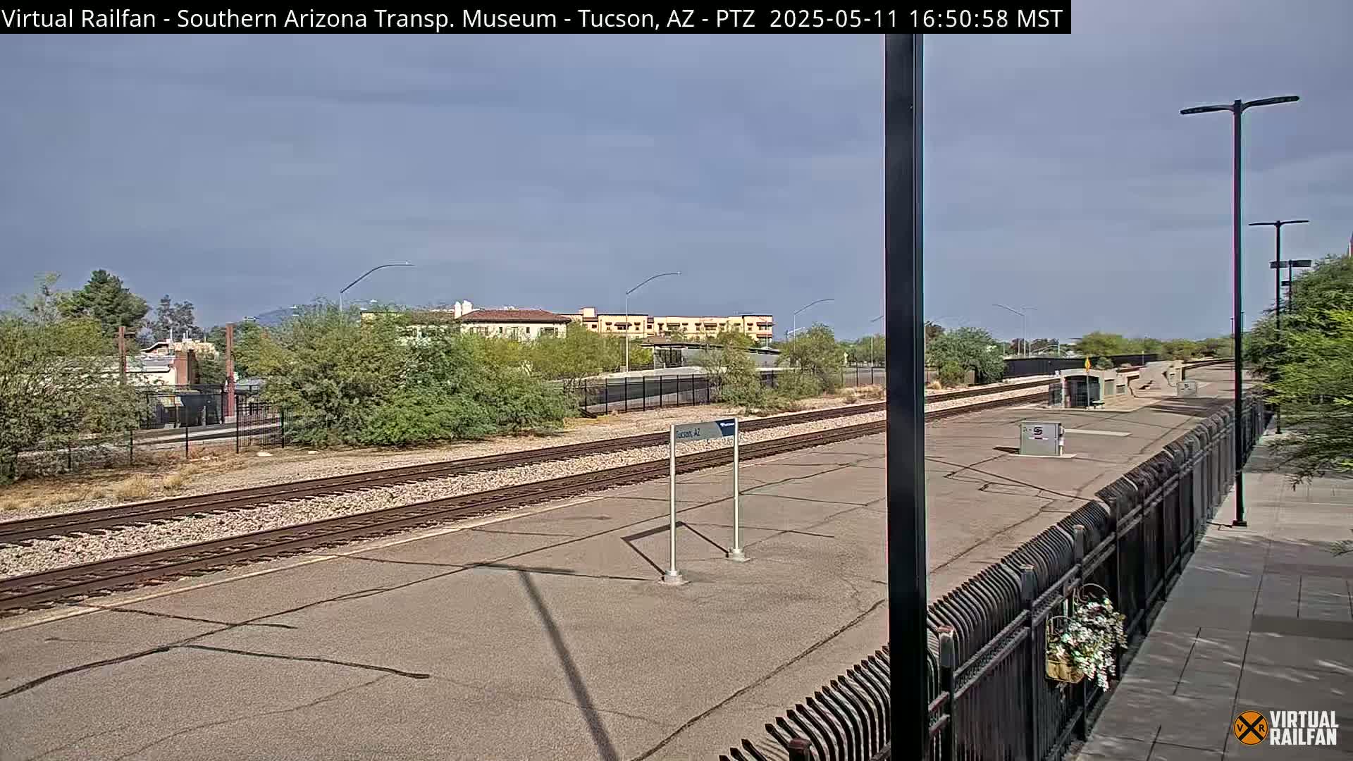 A mostly sunny day reveals train tracks and a platform at a station, with low desert vegetation and buildings in the background.