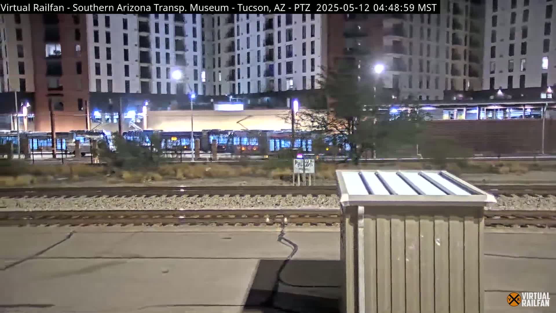 At night, several light rail vehicles are parked near a multi-story building next to train tracks.