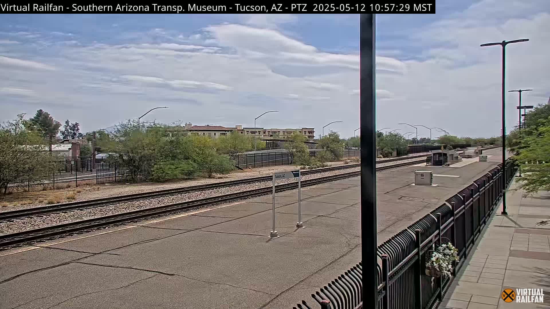 A partly cloudy day reveals a train platform with parallel train tracks, a chain-link fence, and desert landscaping.