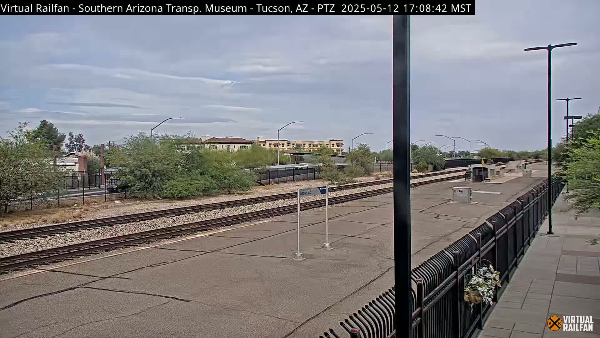 A train station platform with train tracks extending into the distance under a mostly cloudy sky.