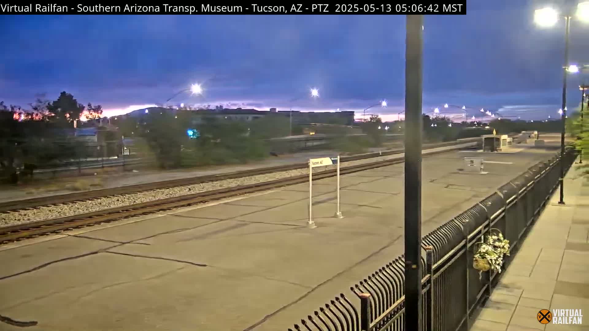 A twilight view of a train station platform with railroad tracks, a fence, and streetlights under a mostly clear sky.