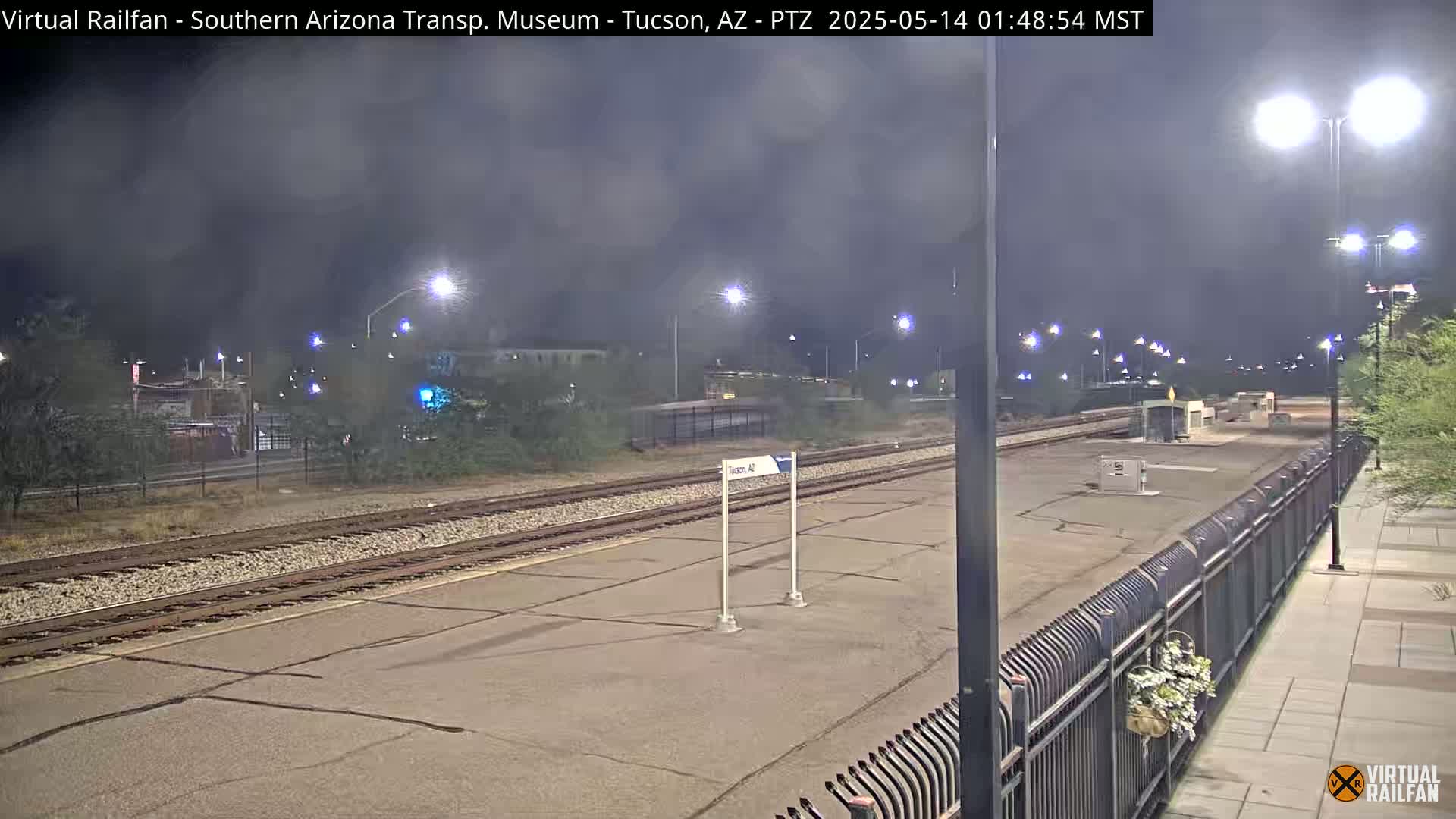 A nighttime view of a train station platform with train tracks, and streetlights under a cloudy sky.