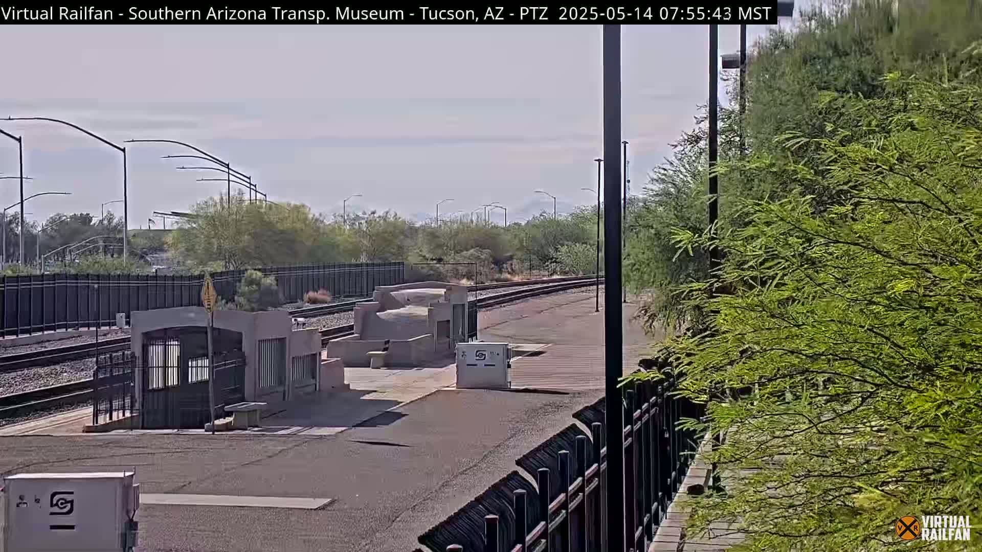 A mostly sunny view of a train station platform with a few small structures and some desert vegetation.